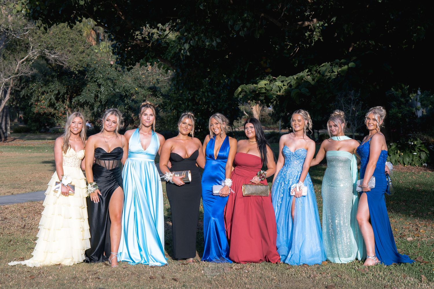 A row of people in blue and coral dresses sitting outdoors on a grassy lawn for a formal event.