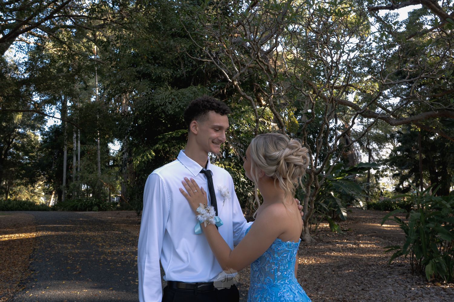 A couple in formal attire dances together on a tree-lined path during an outdoor celebration.