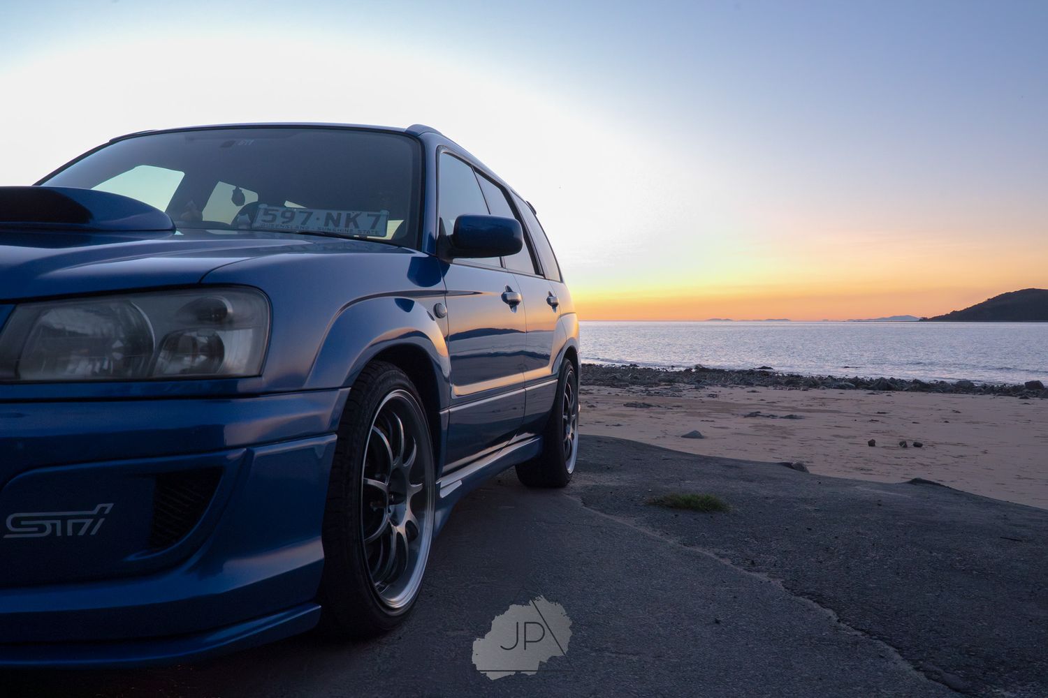 Blue BMW M3 parked on sandy beach at sunset with ocean waves visible in background.