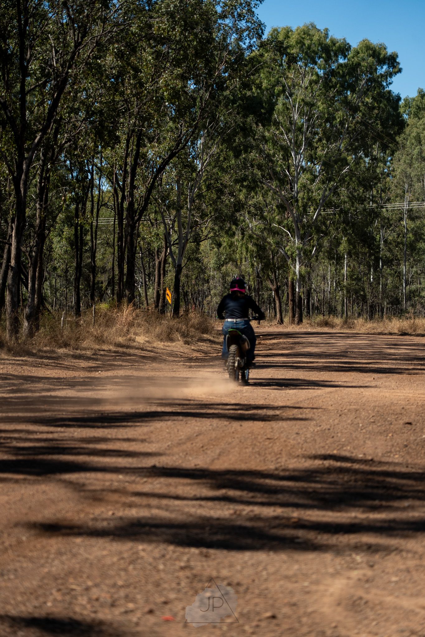 Dirt trail winding through eucalyptus forest with motorcyclists riding in distance on sunny day.