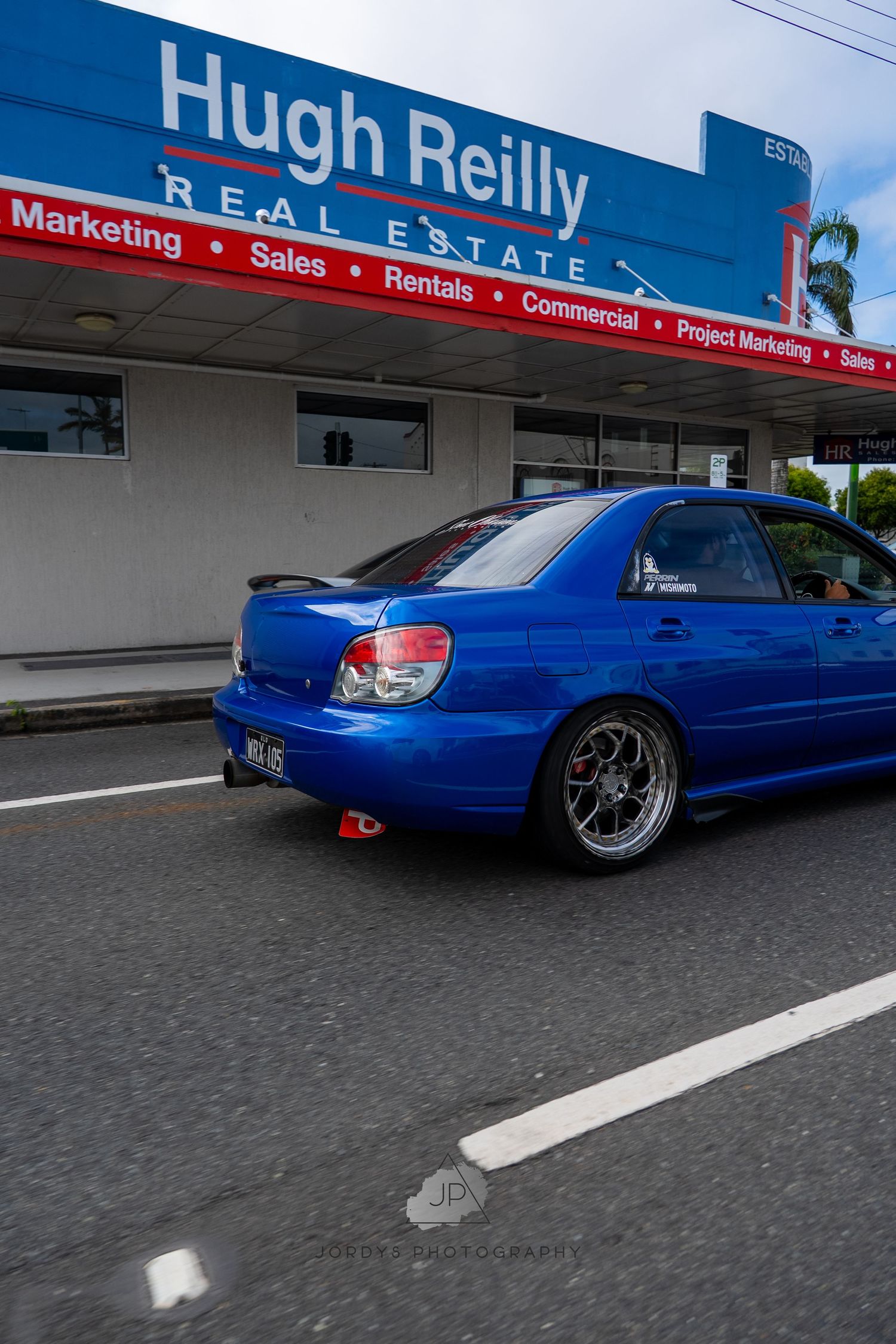 A lowered bright blue Subaru parked outside a real estate office building with custom wheels and sport modifications.