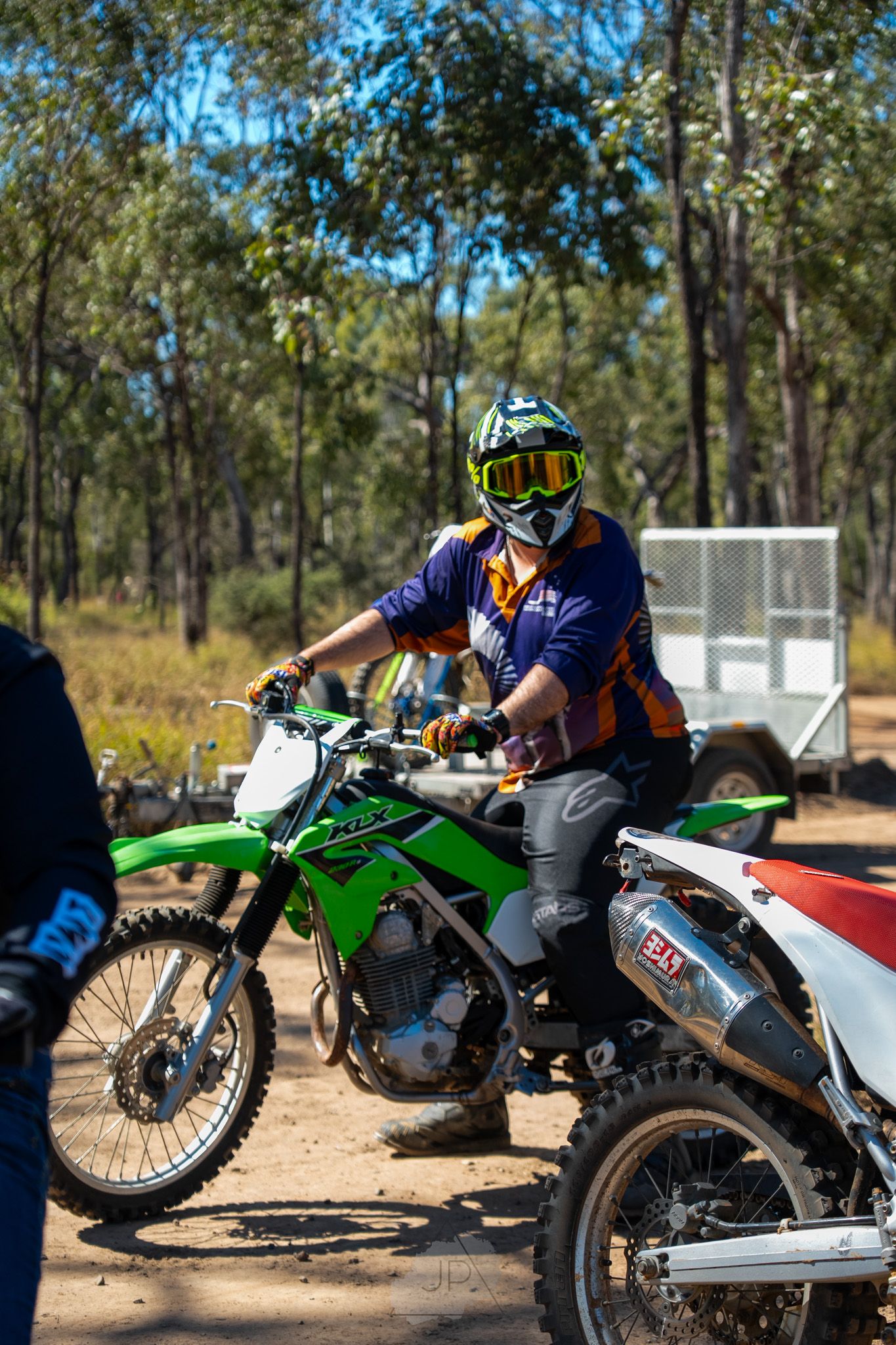 Motorcyclist on a green dirt bike wearing protective gear pauses on a forest trail on a sunny day.