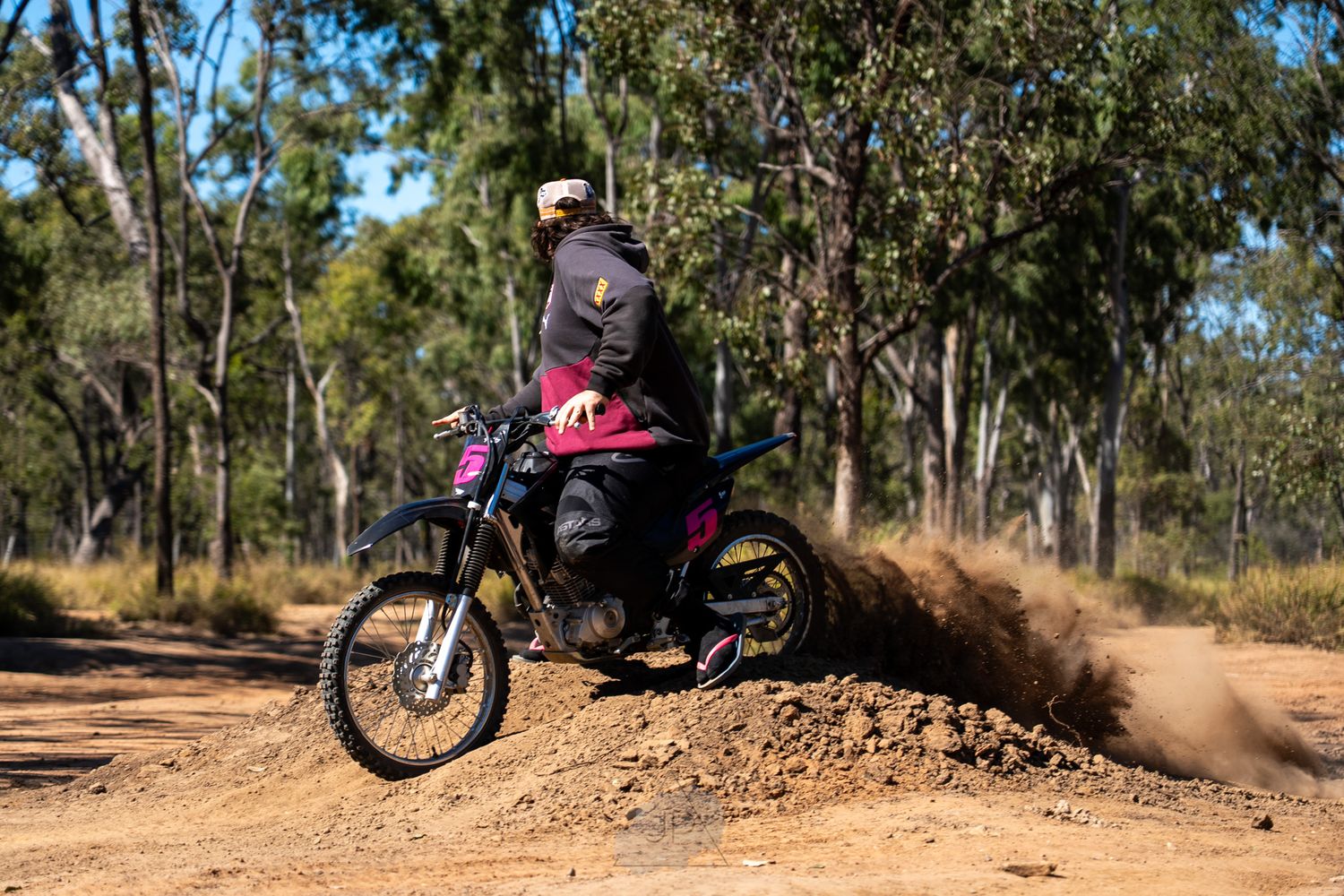 Dirt bike rider kicks up dust while riding through a wooded trail on a red motorcycle in rugged terrain.