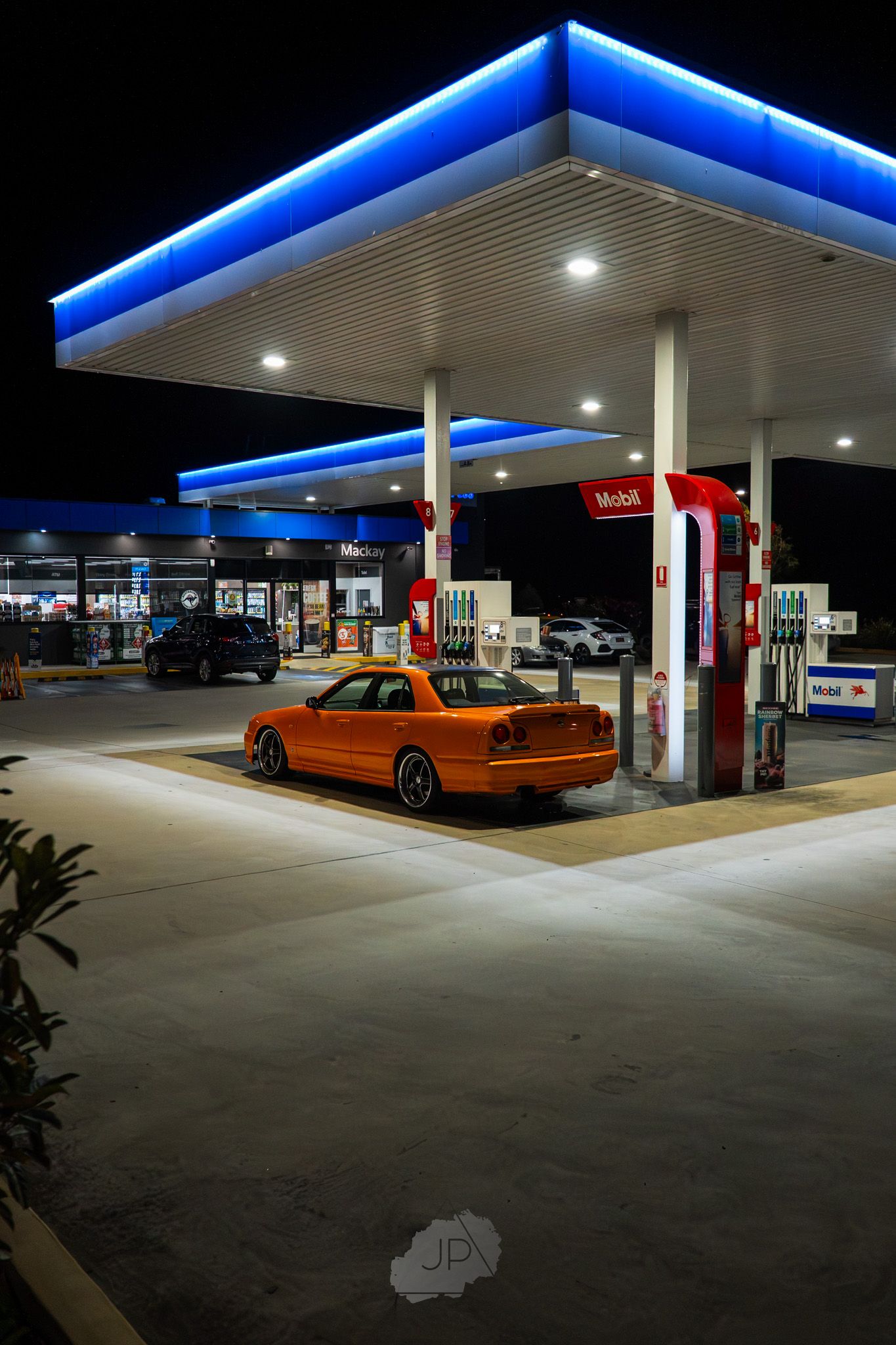 A modern gas station at night illuminated by bright blue LED lighting with an orange car parked at the pump.