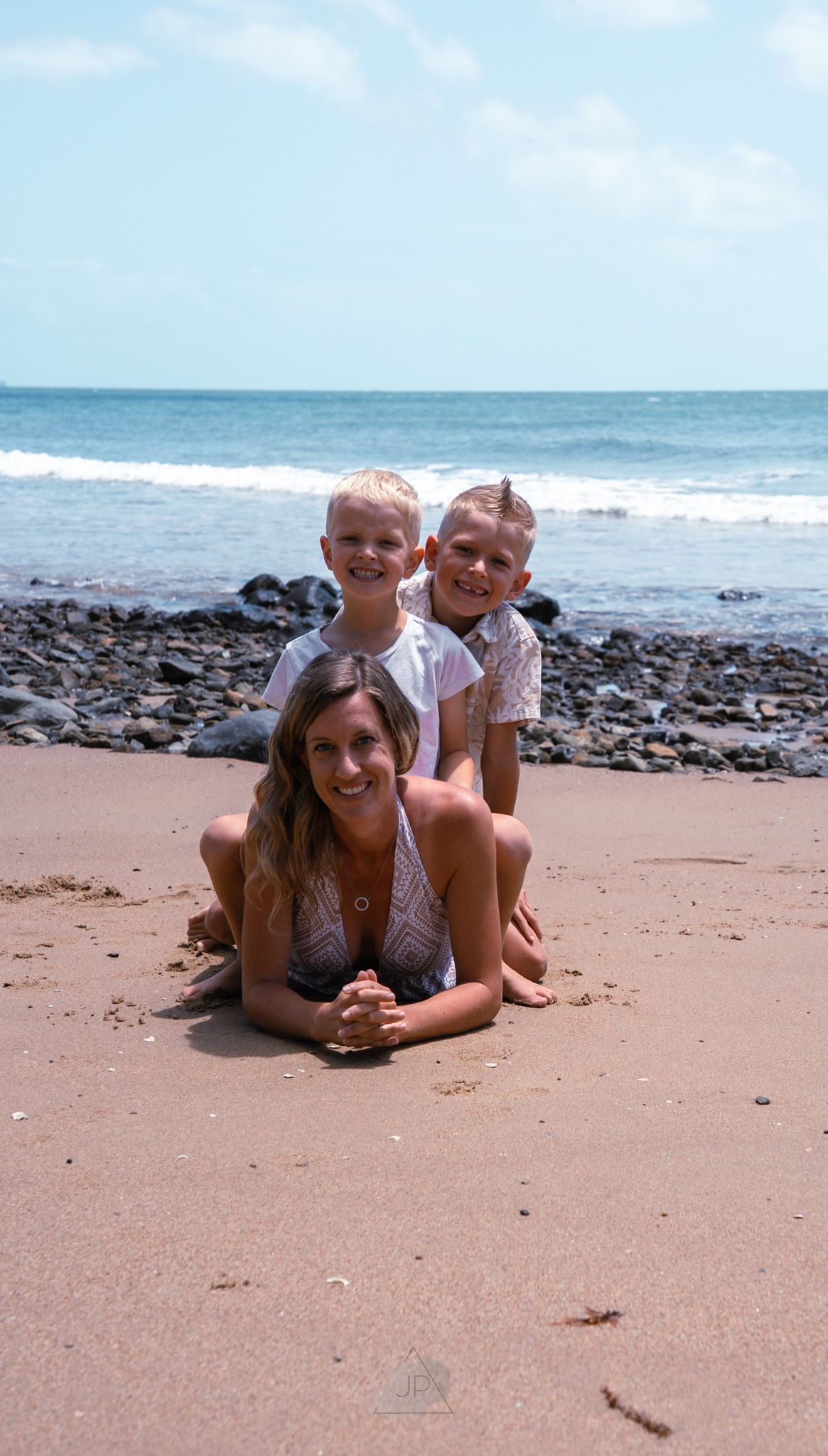 Three people lying on a sandy beach with rocks and ocean waves in the background.