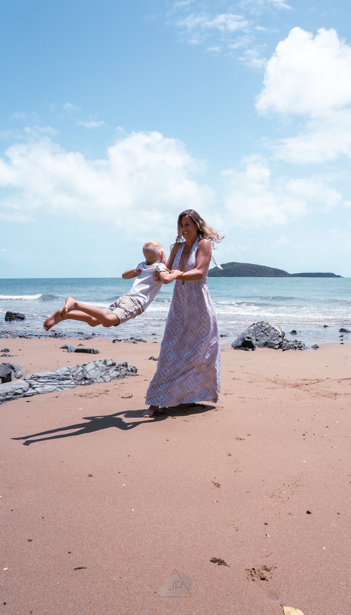 A person in a white dress walks along a scenic tropical beach with rocky outcrops and blue ocean views.