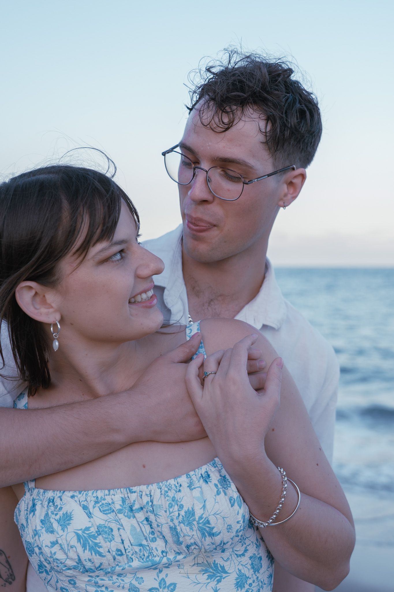 A romantic couple embraces at sunset by the ocean, with the woman wearing a blue and white floral dress.