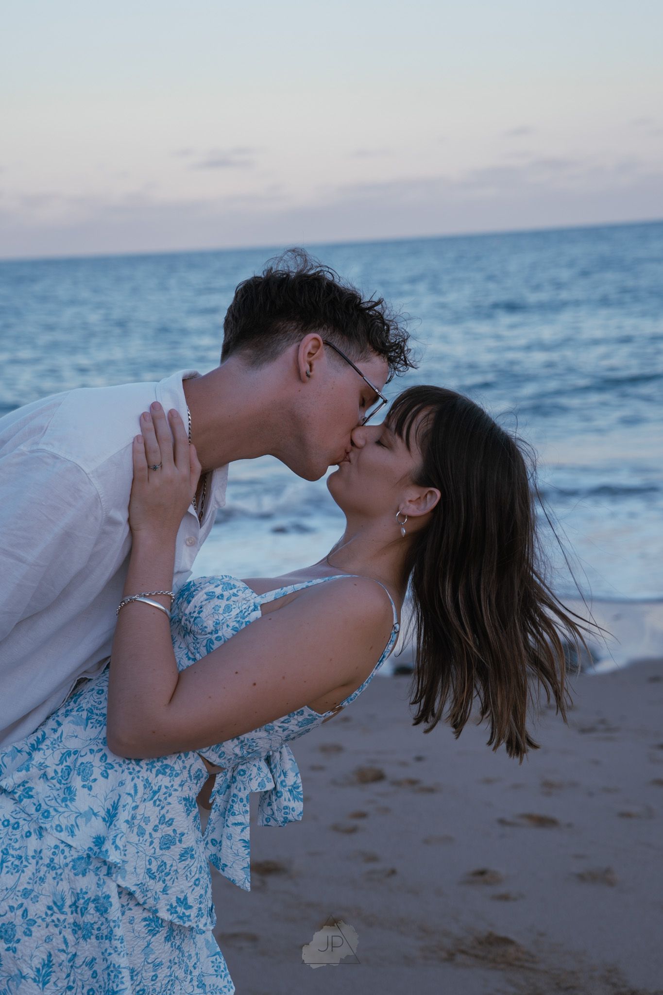 A couple shares a romantic kiss on the beach at sunset with ocean waves in the background.