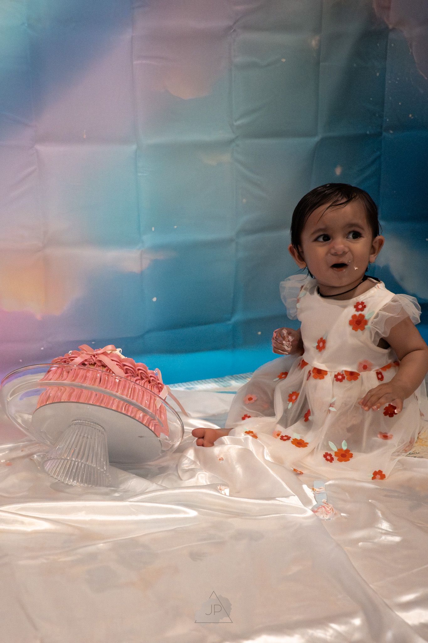 A baby wearing a white floral dress lays on a white surface in a dreamy pastel-colored setting.