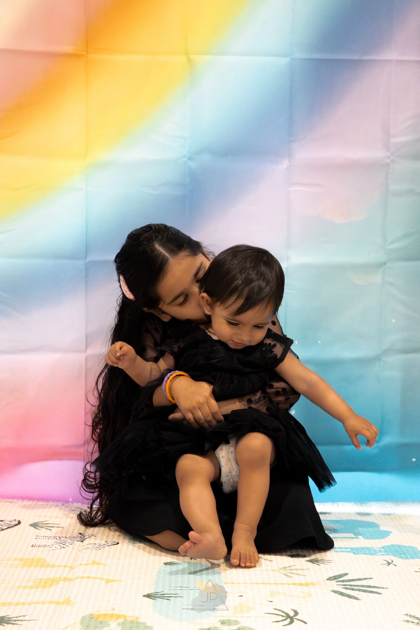 Two people in black outfits sit close together on a colorful surface with soft pastel lighting in the background.