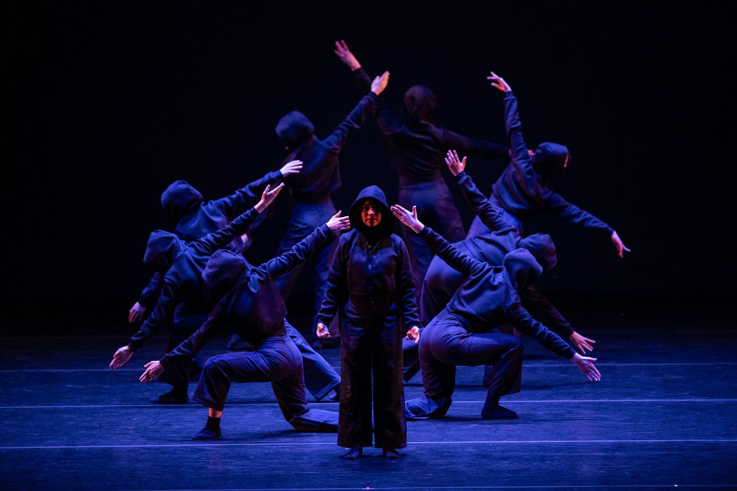 Dancers in blue costumes perform a synchronized modern dance formation around a central figure on a dark stage.