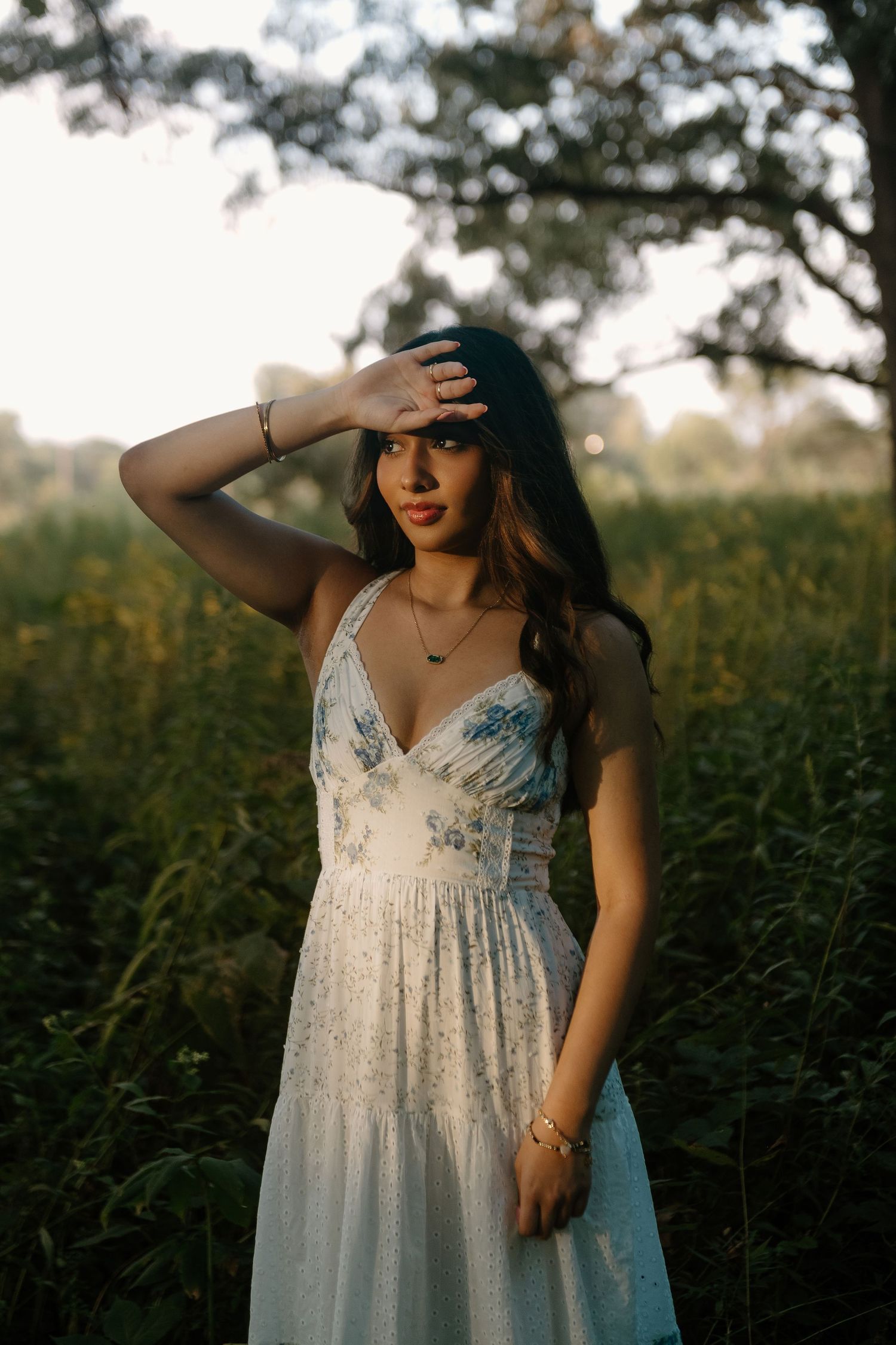 Woman in a white floral sundress shields her eyes from the sun while standing in a golden-lit meadow beneath tall trees.