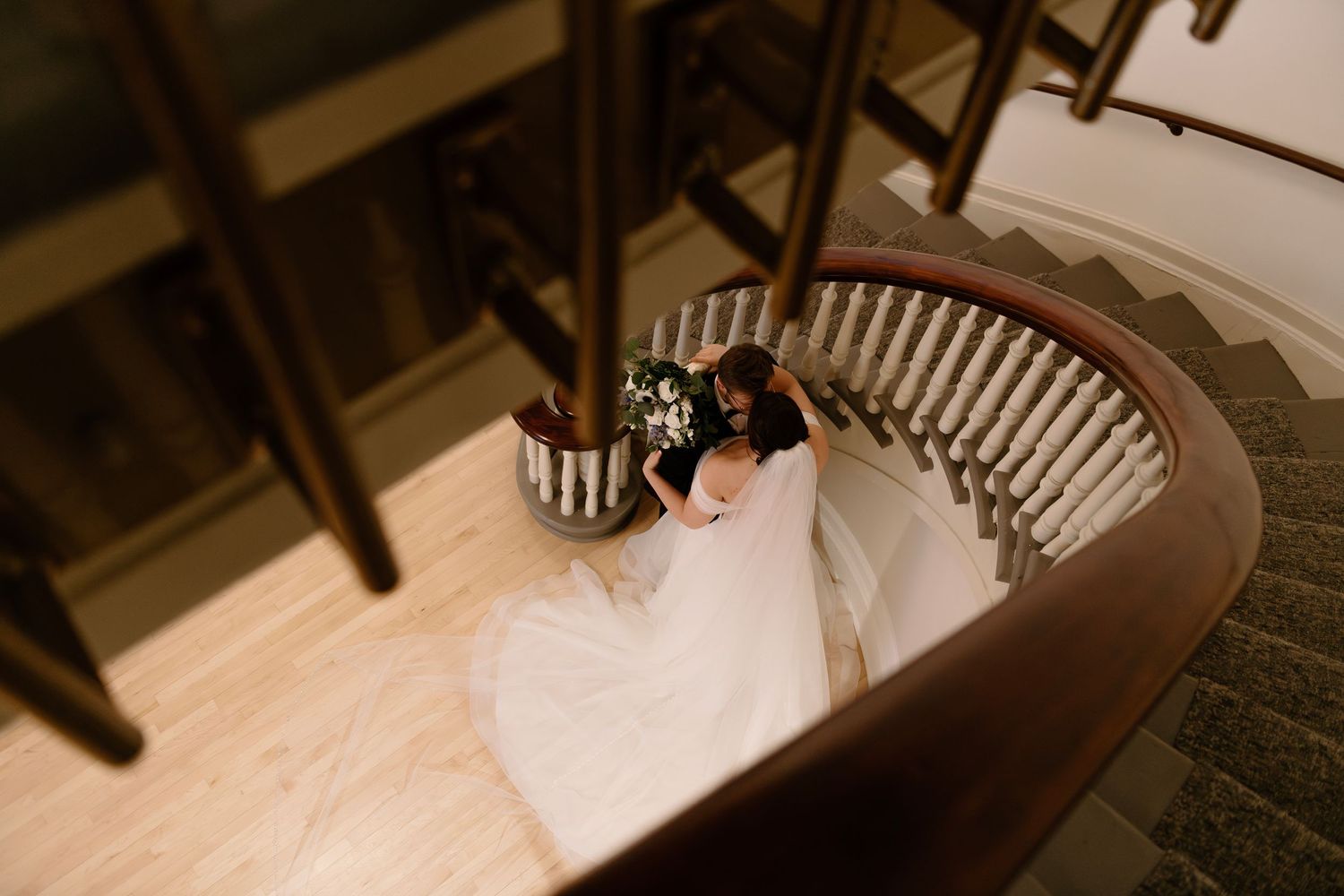 Aerial view of a bride in a white gown holding a bouquet on an elegant curved staircase.