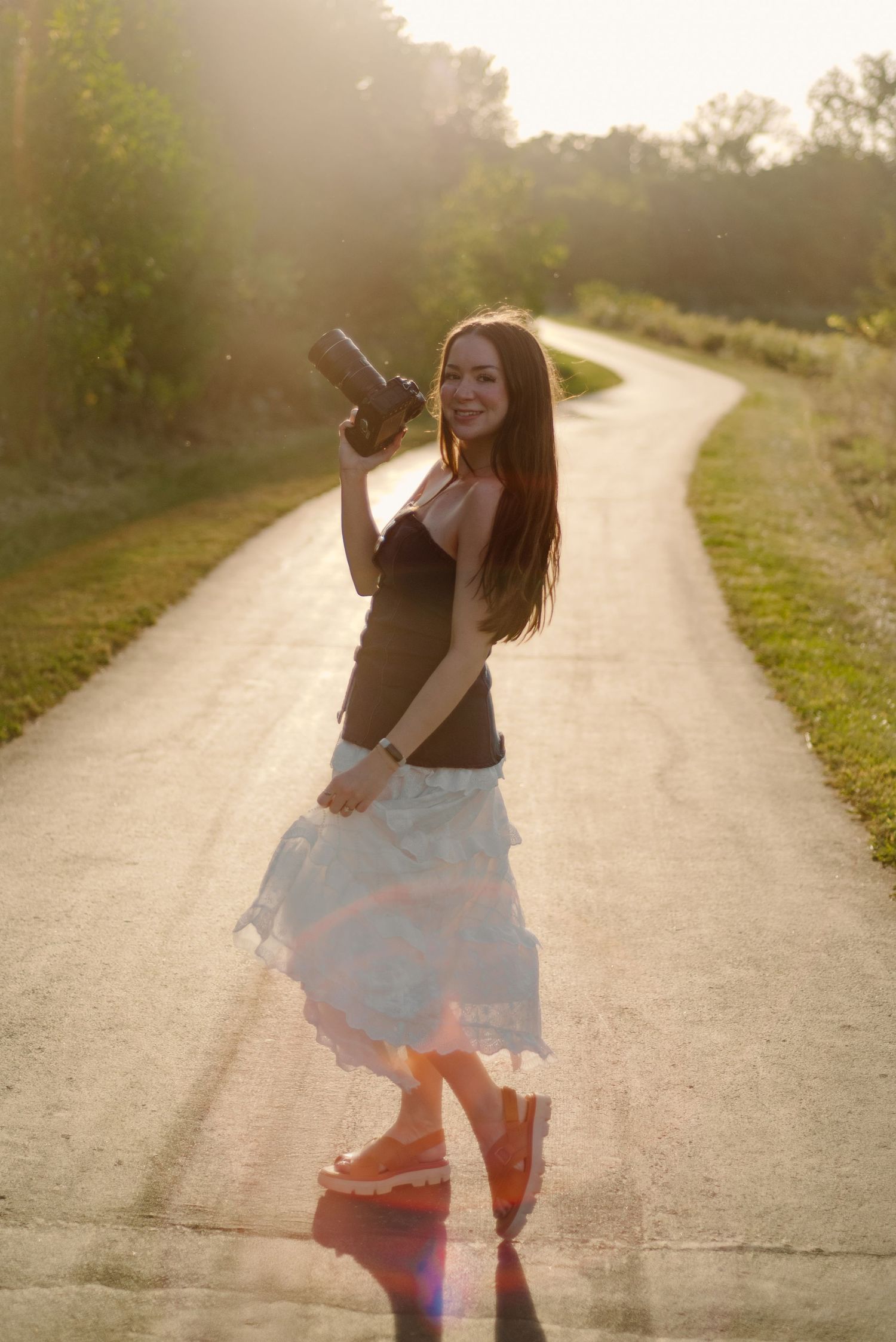 Young PHOTOGRAPHER woman in white skirt holds camera on sunny winding road, golden backlight illuminating her smile.