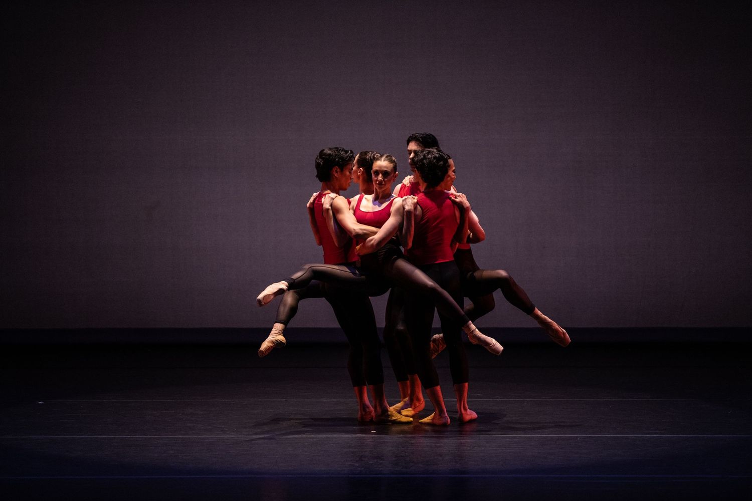 Modern dancers in red costumes perform an expressive group movement on a dark stage.
