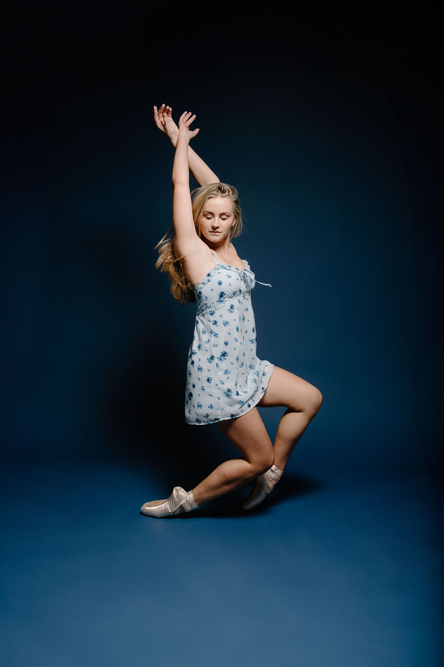 Blonde woman in floral blue dress dancing gracefully with arms raised on dark blue studio background.