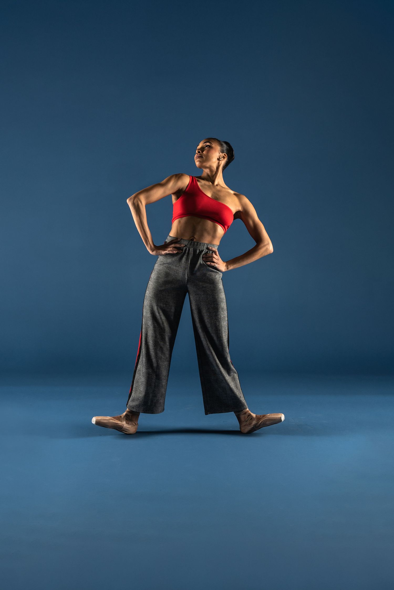 Athletic dancer in red top and gray pants performs dynamic lunging movements against blue studio backdrop.