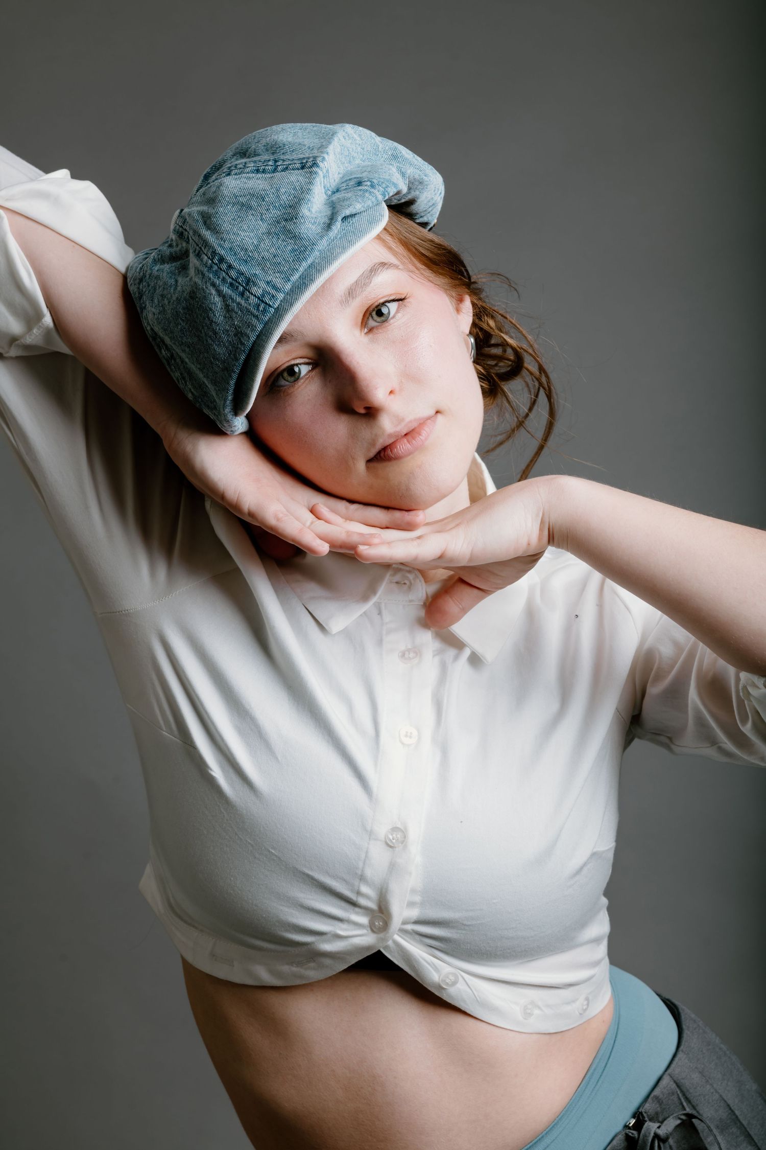 Figure in a denim cap and white tied shirt poses artistically against gray backdrop in a studio portrait setting.
