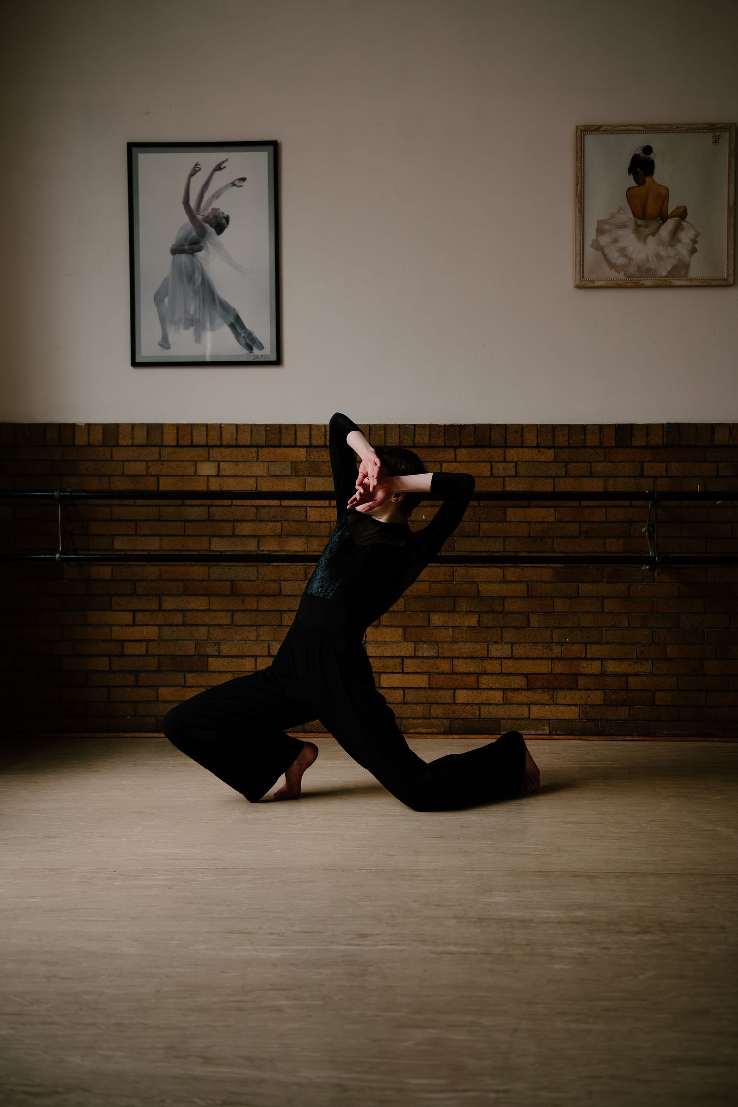 A dancer in a black outfit performs an expressive movement against a brick wall with framed artwork in a dance studio.