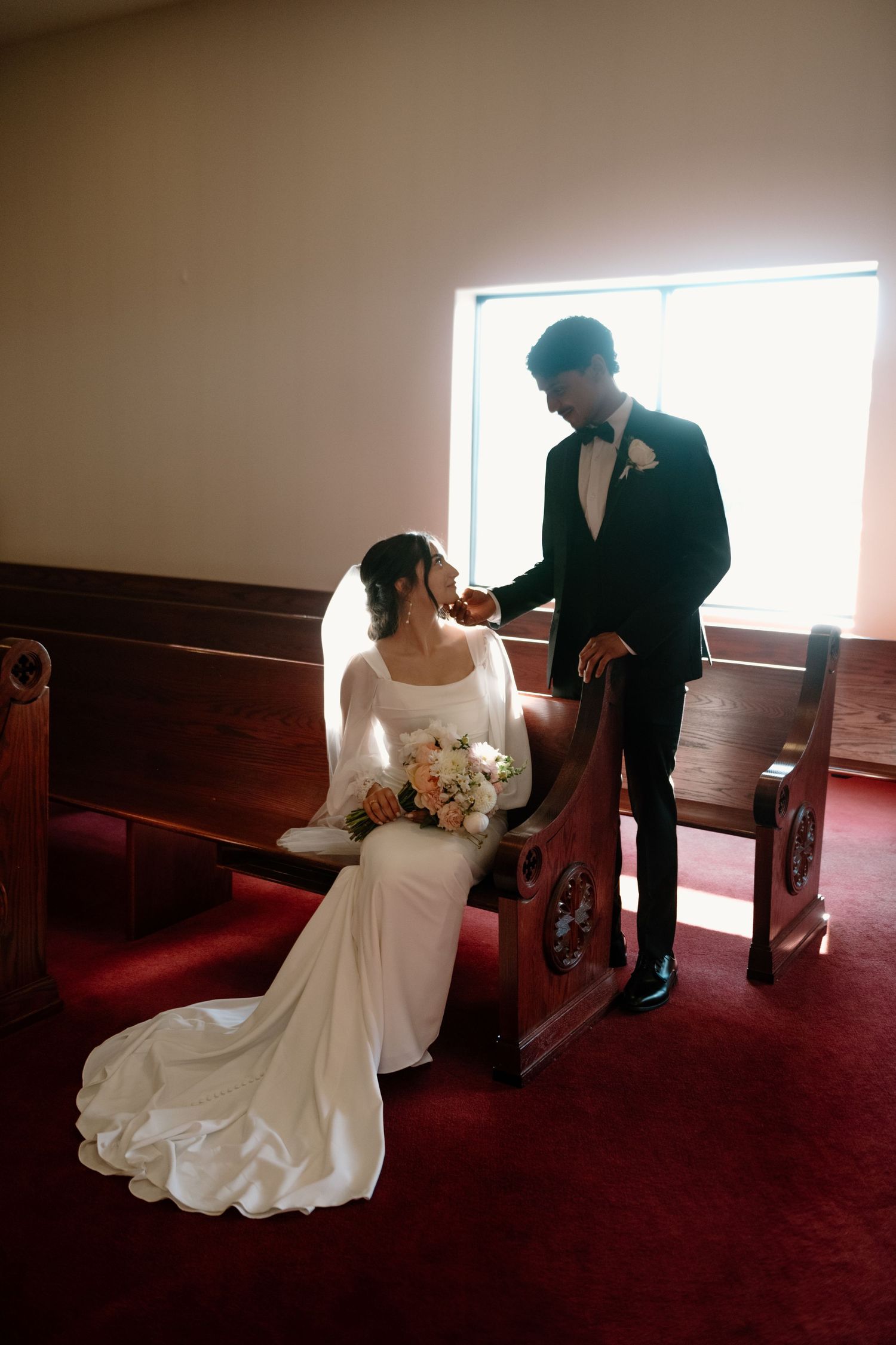 A couple shares an intimate moment in a church with red carpeting, the bride wearing a long-sleeved white gown holding flowers.