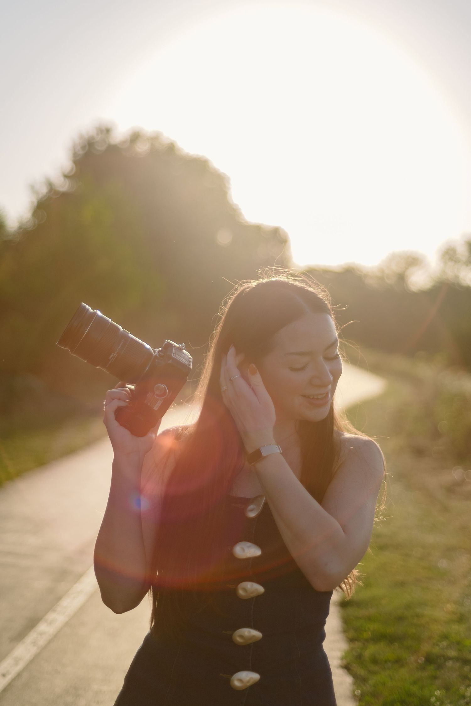 Woman PHOTOGRAPHER in black dress holding DSLR camera outdoors during golden hour with sun flare in background.