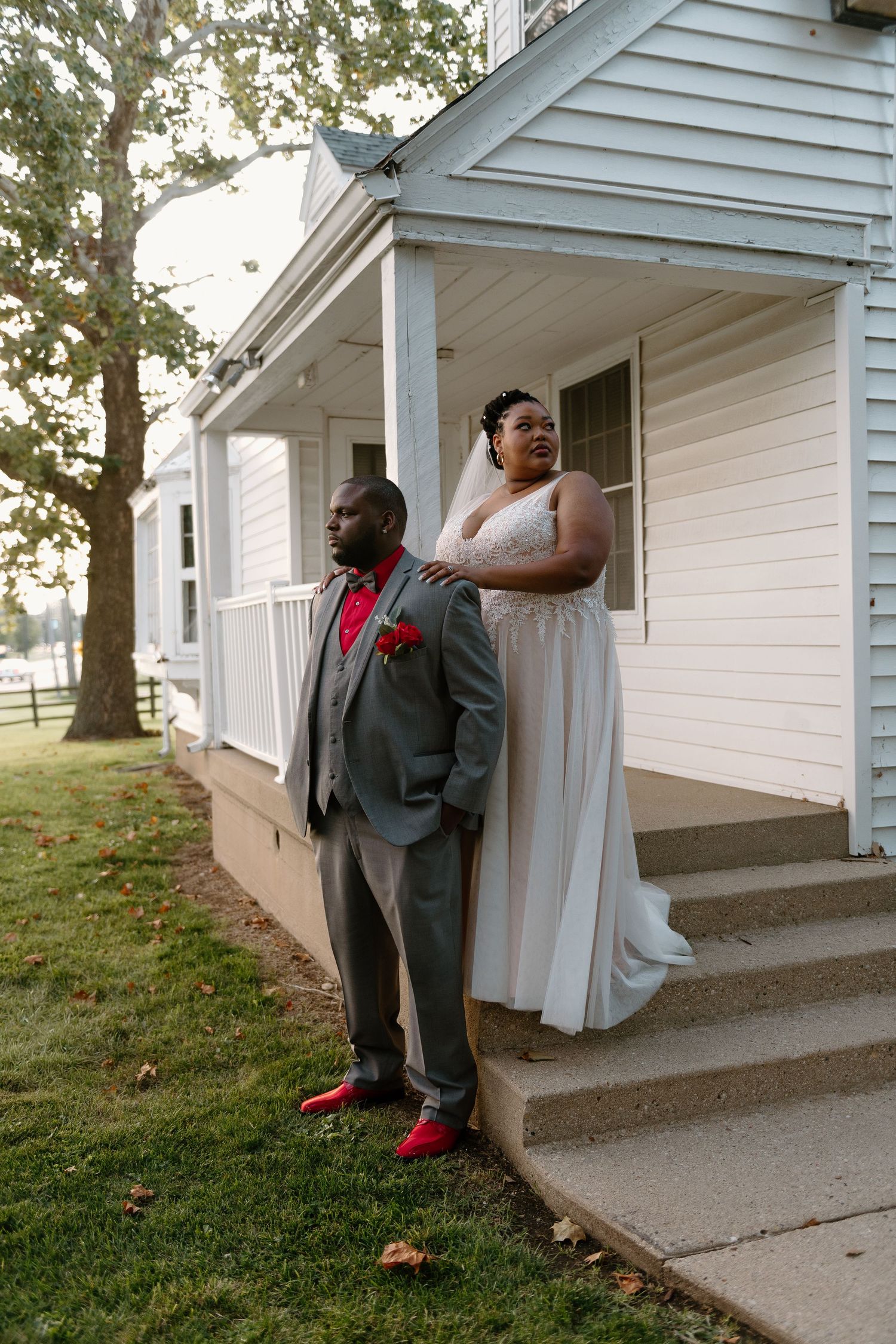 A couple shares a tender moment on the steps of a white house, with the groom wearing a gray suit and red shoes.