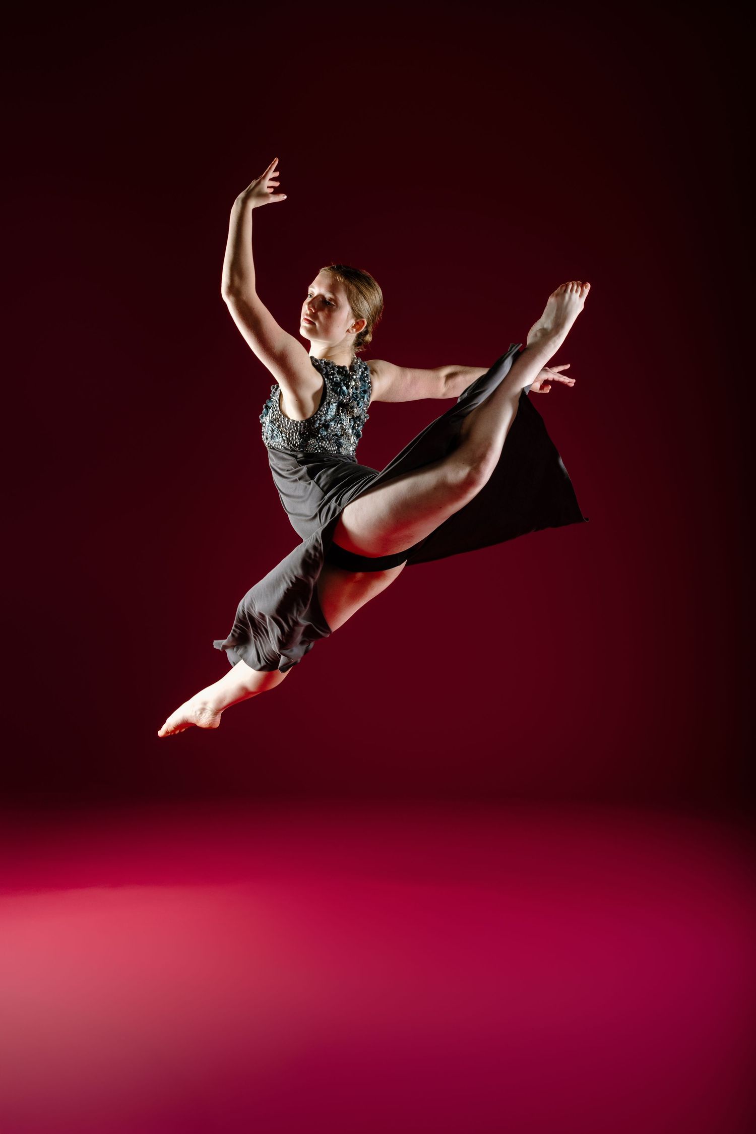 Dancer performs graceful leap in gray dress against dark red background with dramatic lighting and elegant form.