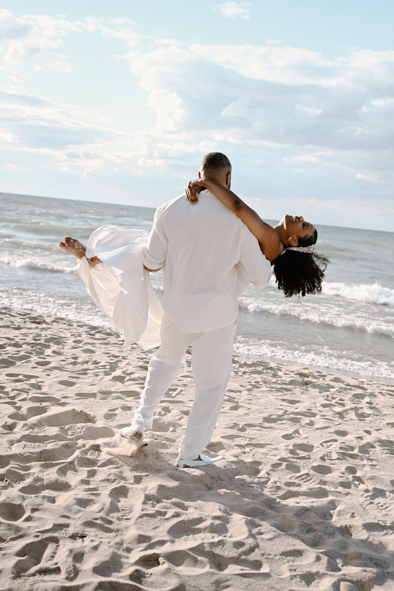 A couple in matching white outfits performs dance moves together on a sunny beach with ocean waves in background.