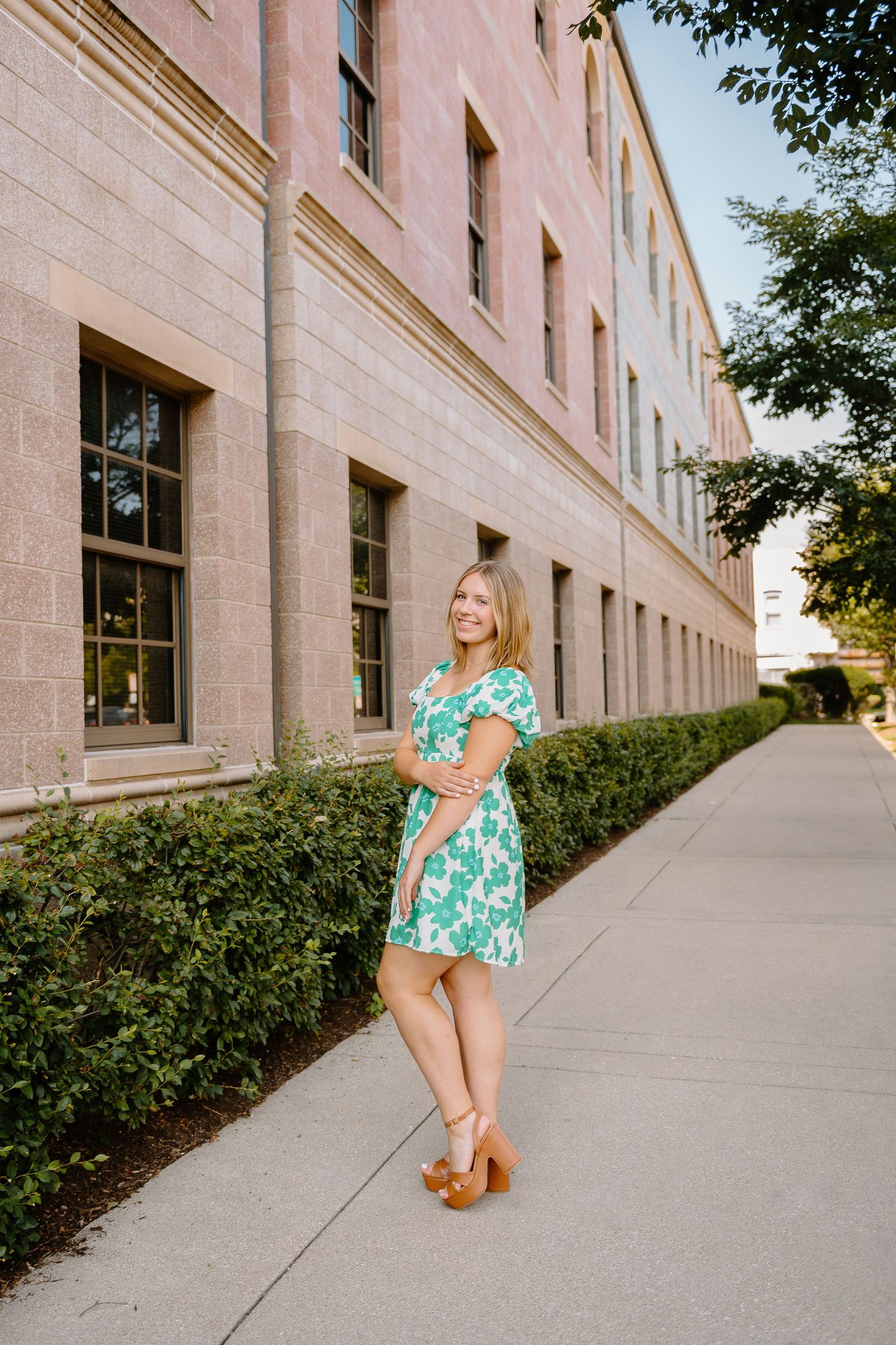 Smiling woman in a green floral dress and tan platform sandals poses on a sidewalk beside a pink brick building and trimmed green hedge.