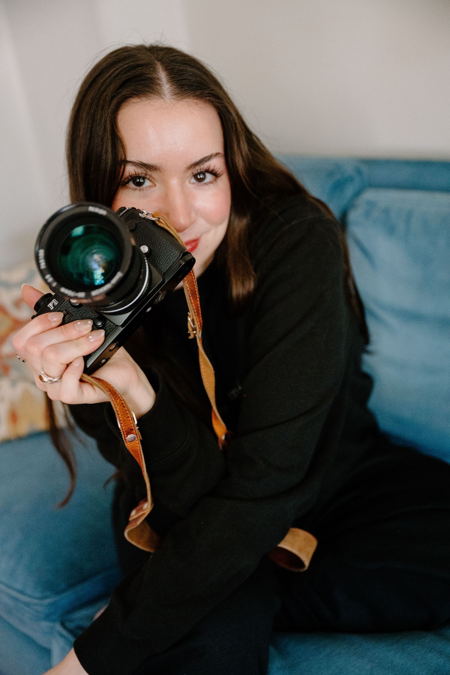 Young PHOTOGRAPHER woman in black outfit holding a vintage film camera on a blue sofa, looking into the lens.
