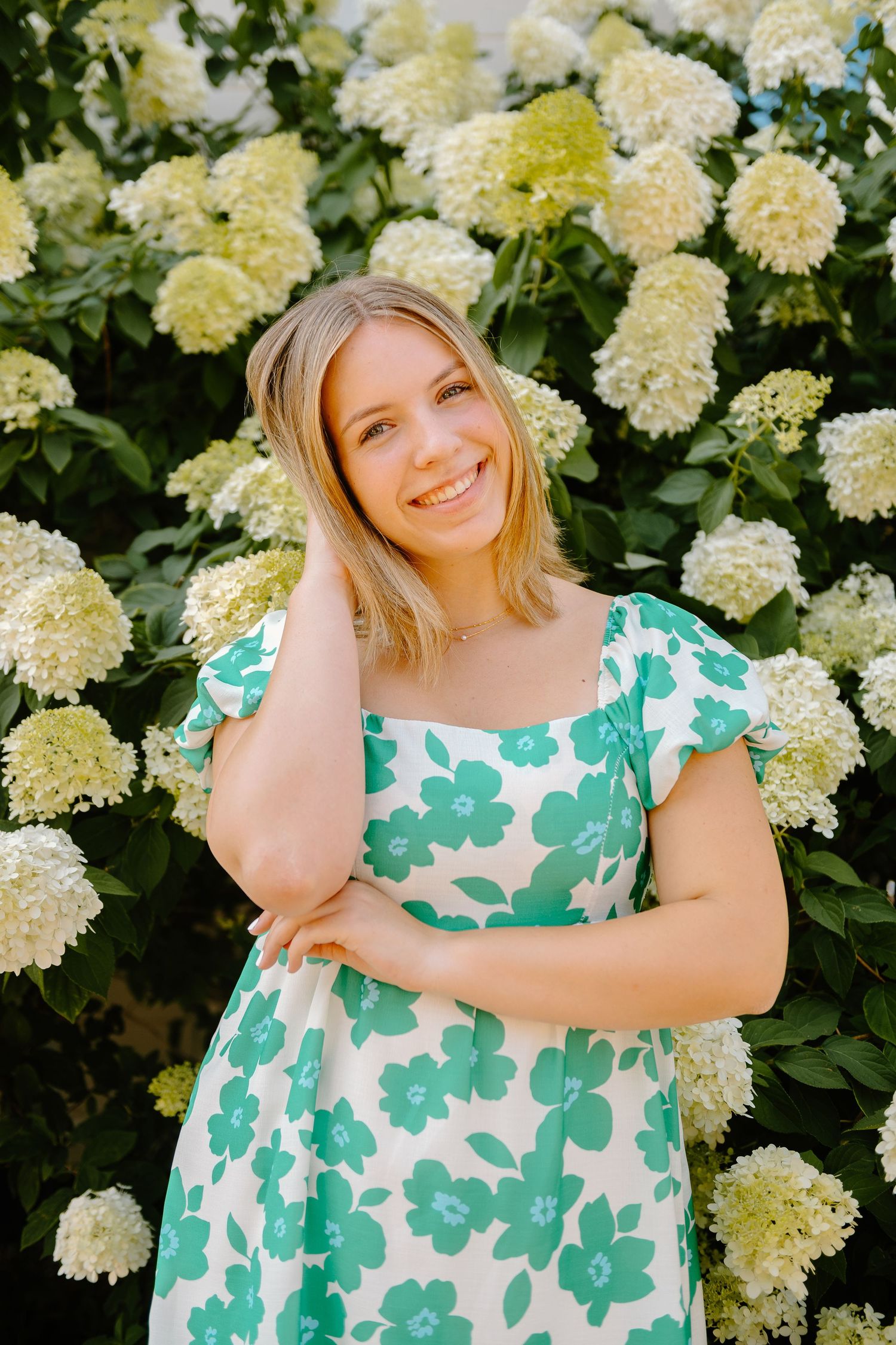 Woman in green and white floral dress poses by blooming white hydrangea bush in garden.