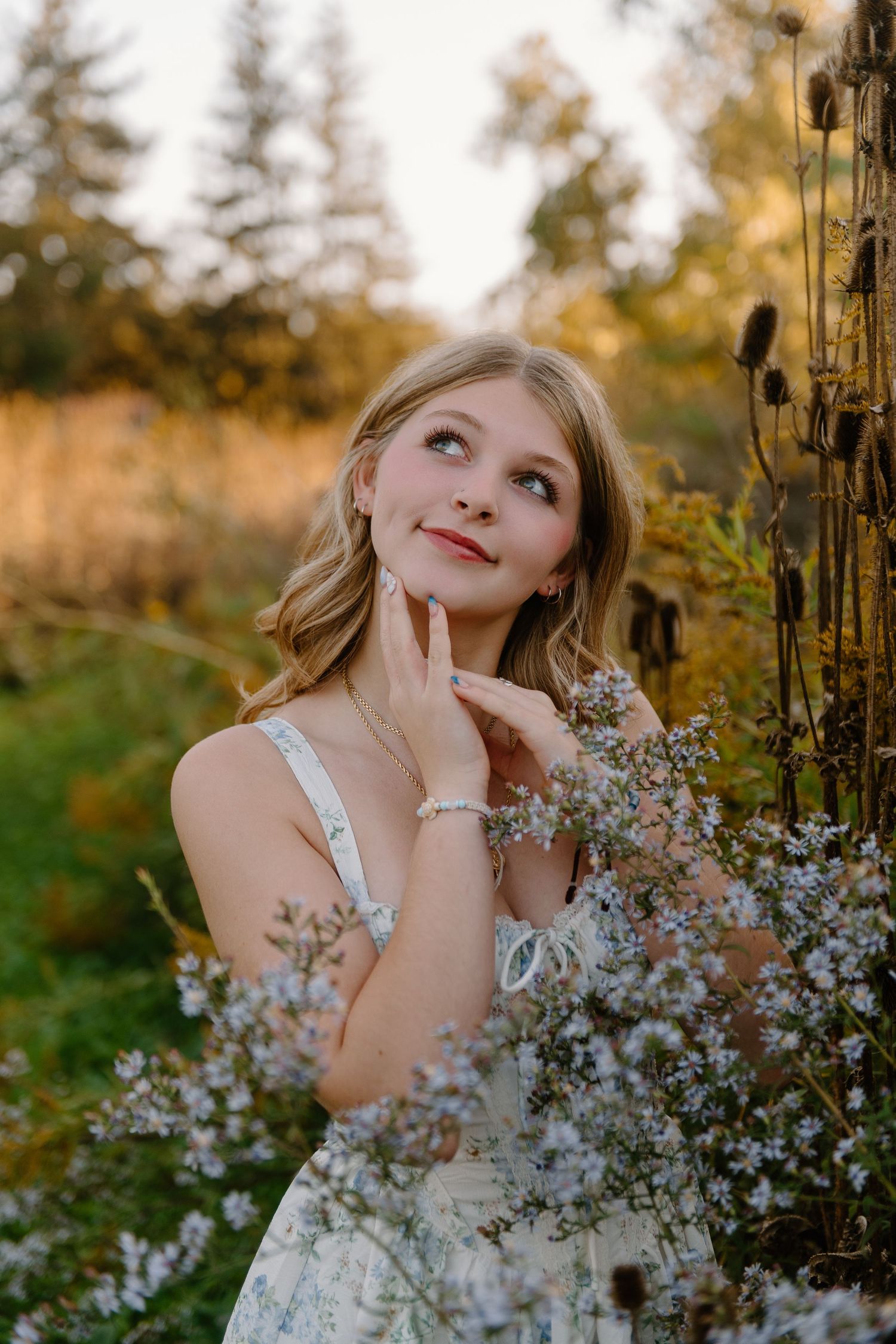 Young blonde woman in floral dress poses dreamily among wildflowers at golden hour in a lush outdoor setting.