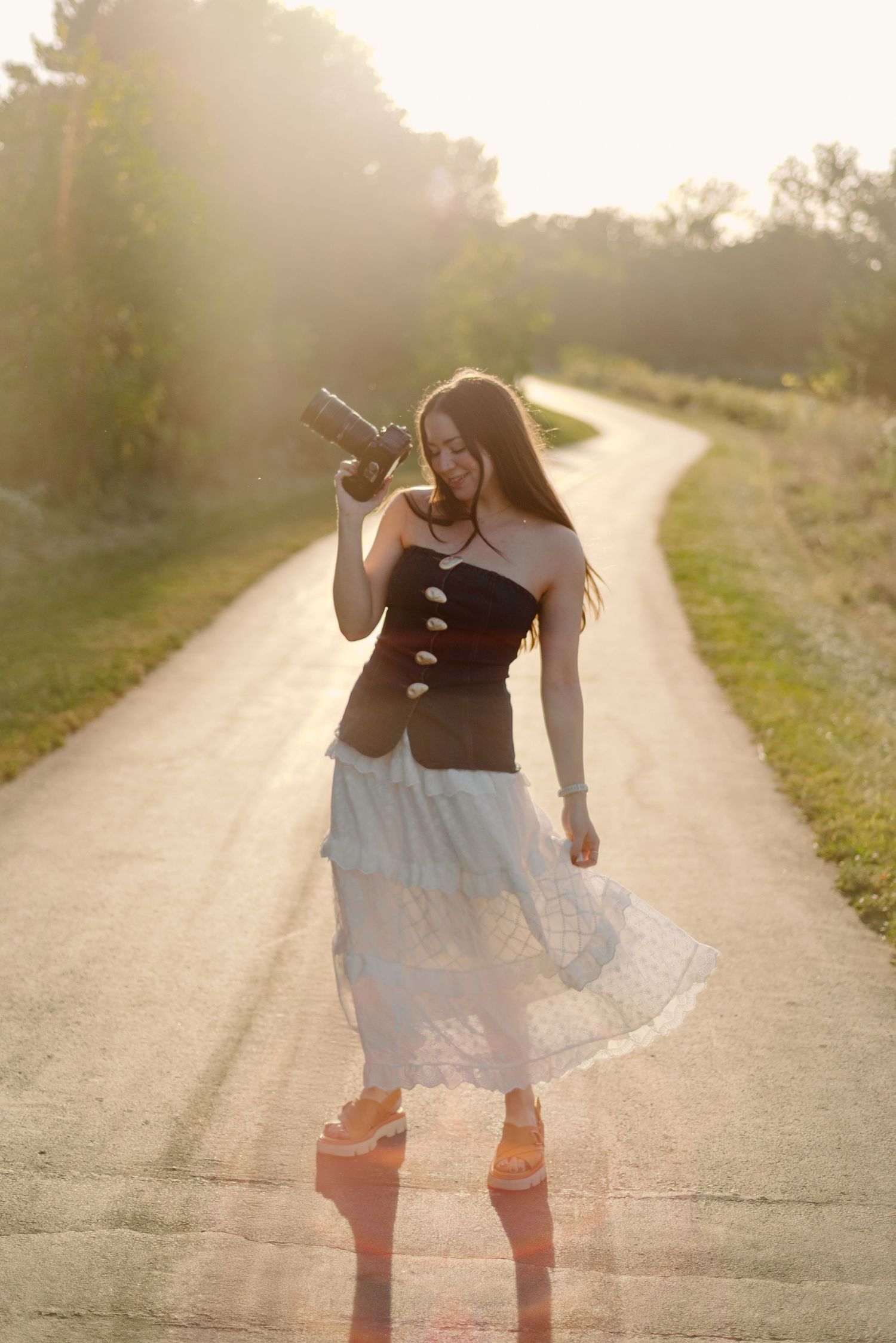 Young PHOTOGRAPHER woman in white skirt holds camera on sunny rural road at golden hour.