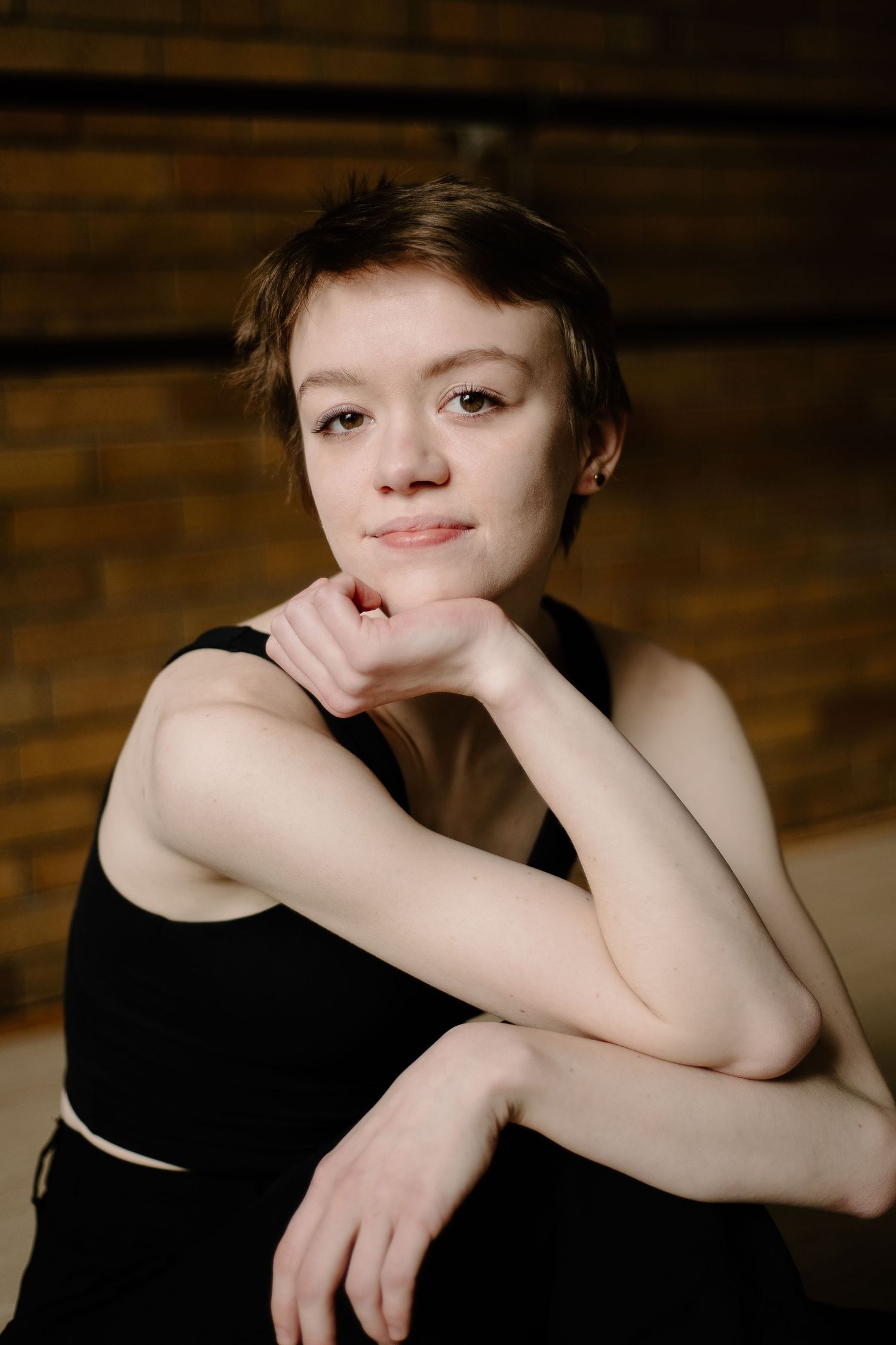 Portrait of a person in a black sleeveless top resting their chin on their hand, photographed against a wooden backdrop.