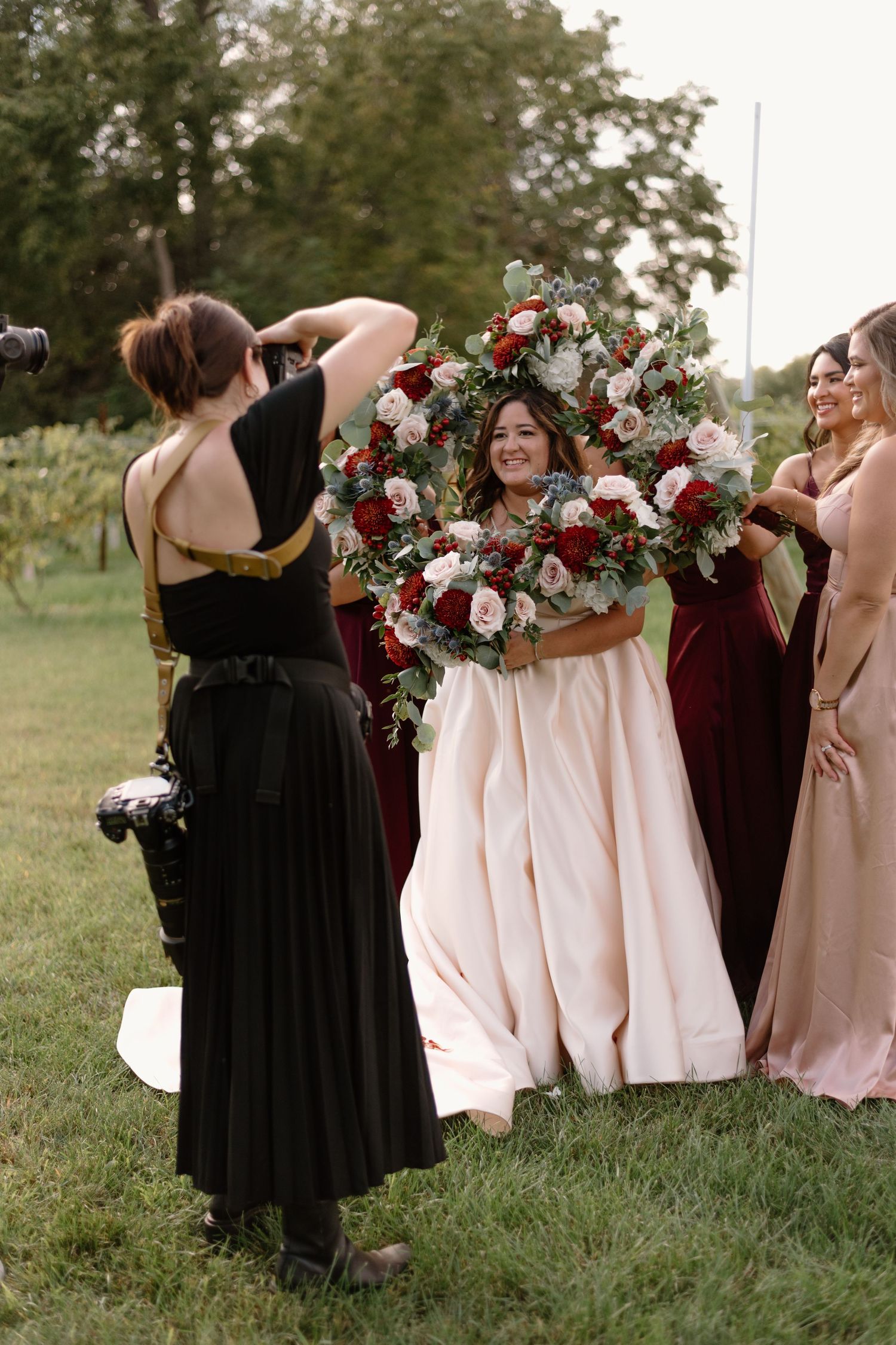 Bridesmaids in burgundy and blush dresses pose together holding floral bouquets in an outdoor garden setting.