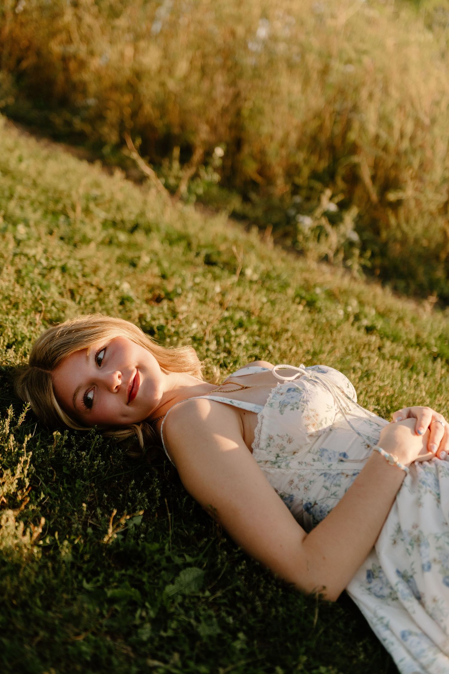 A young blonde woman in a floral sundress lying on green grass, smiling softly in warm golden sunlight.