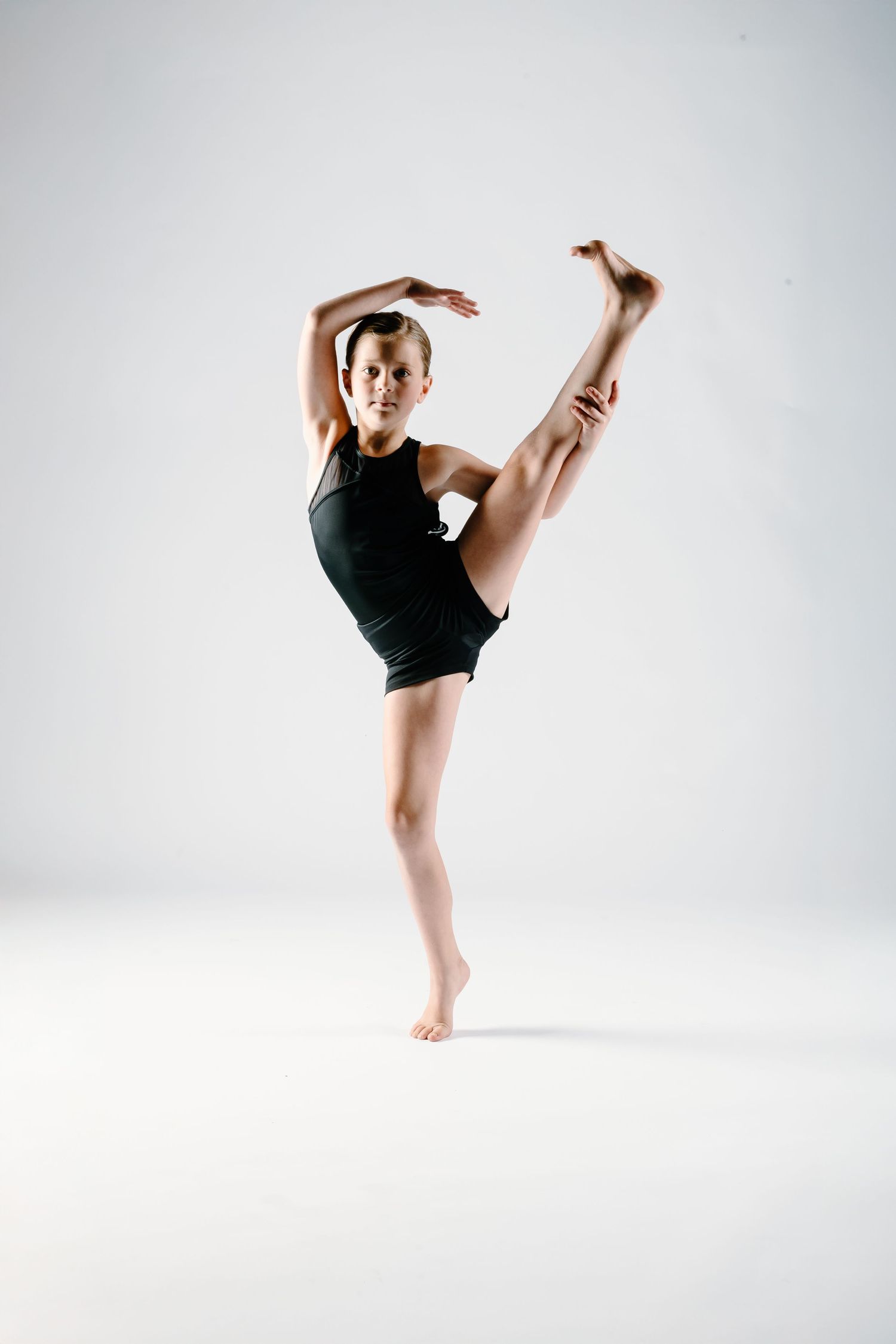 Contemporary dancer in black leotard performs graceful high kick pose on white background.