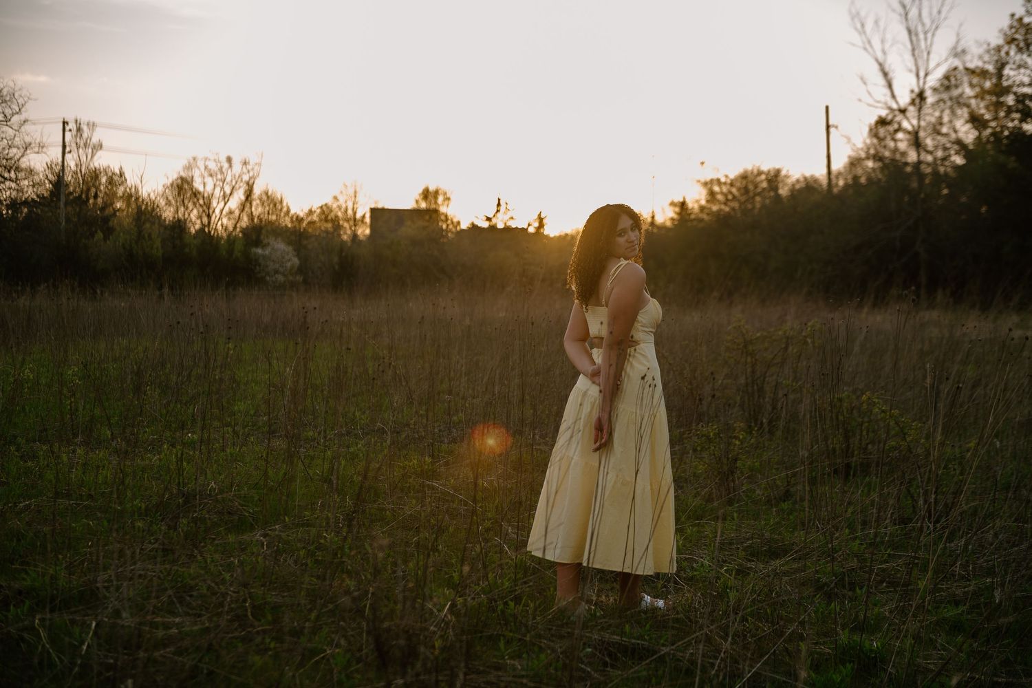 Woman in yellow dress standing in golden hour sunlit field surrounded by tall grass and trees at sunset.