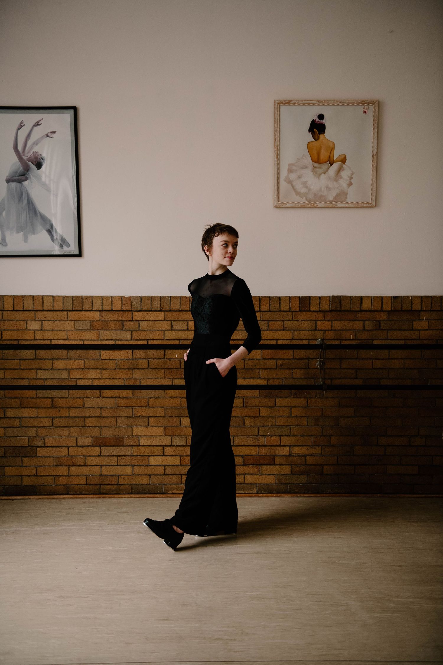 Figure in an elegant black dress poses in front of a brick wall decorated with framed artwork in a minimalist setting.