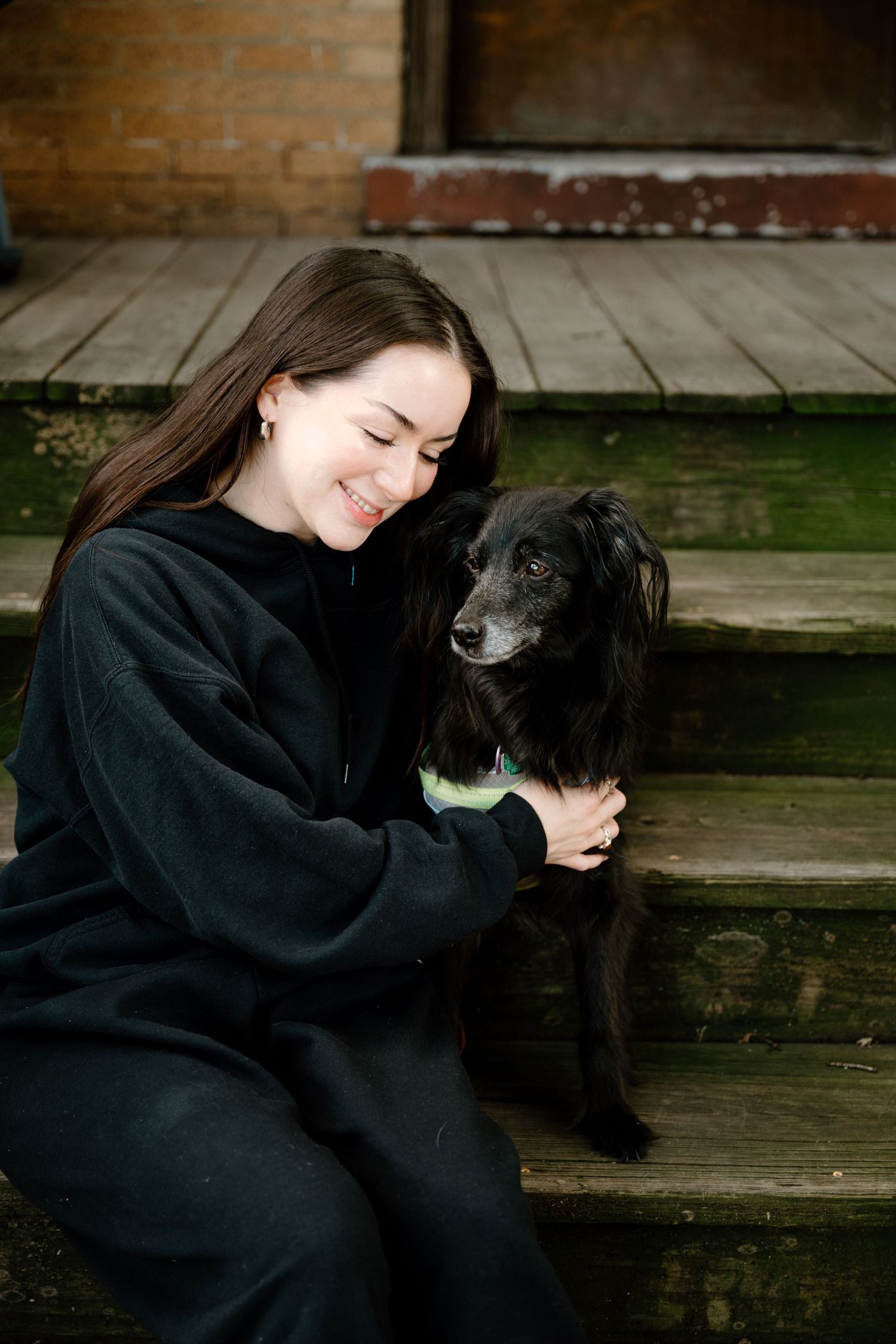 Young PHOTOGRAPHER in black outfit smiling and hugging her black dog while sitting on wooden steps outdoors.