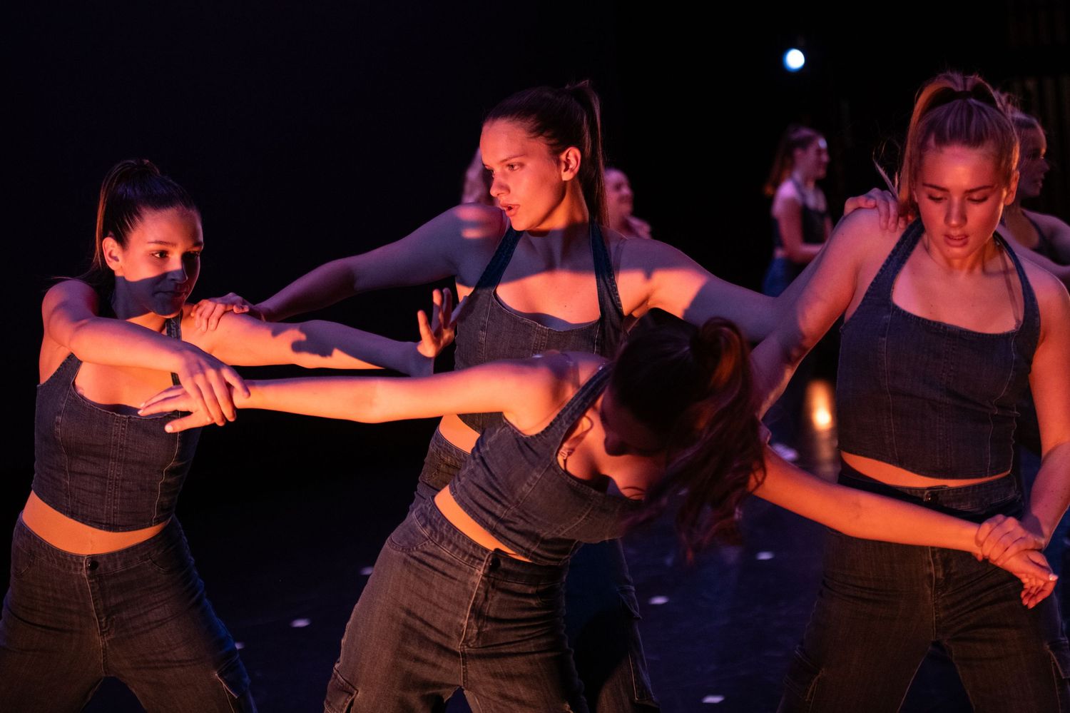 Female dancers perform an intense contemporary routine on a dark stage, wearing matching dark crop tops and jeans.