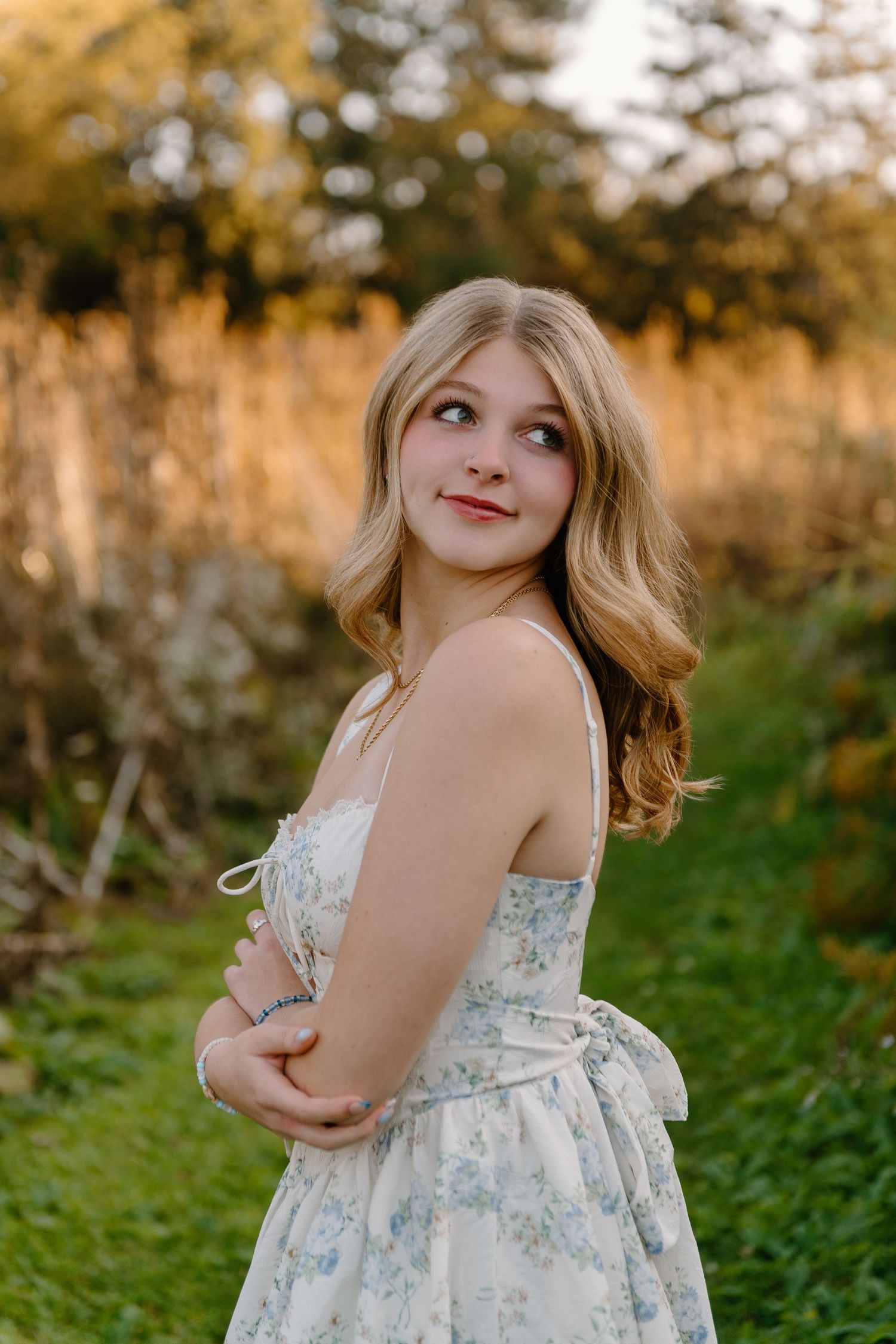 Young woman with wavy blonde hair in a blue floral sundress poses with folded arms and a soft smile in a sunlit grassy field.
