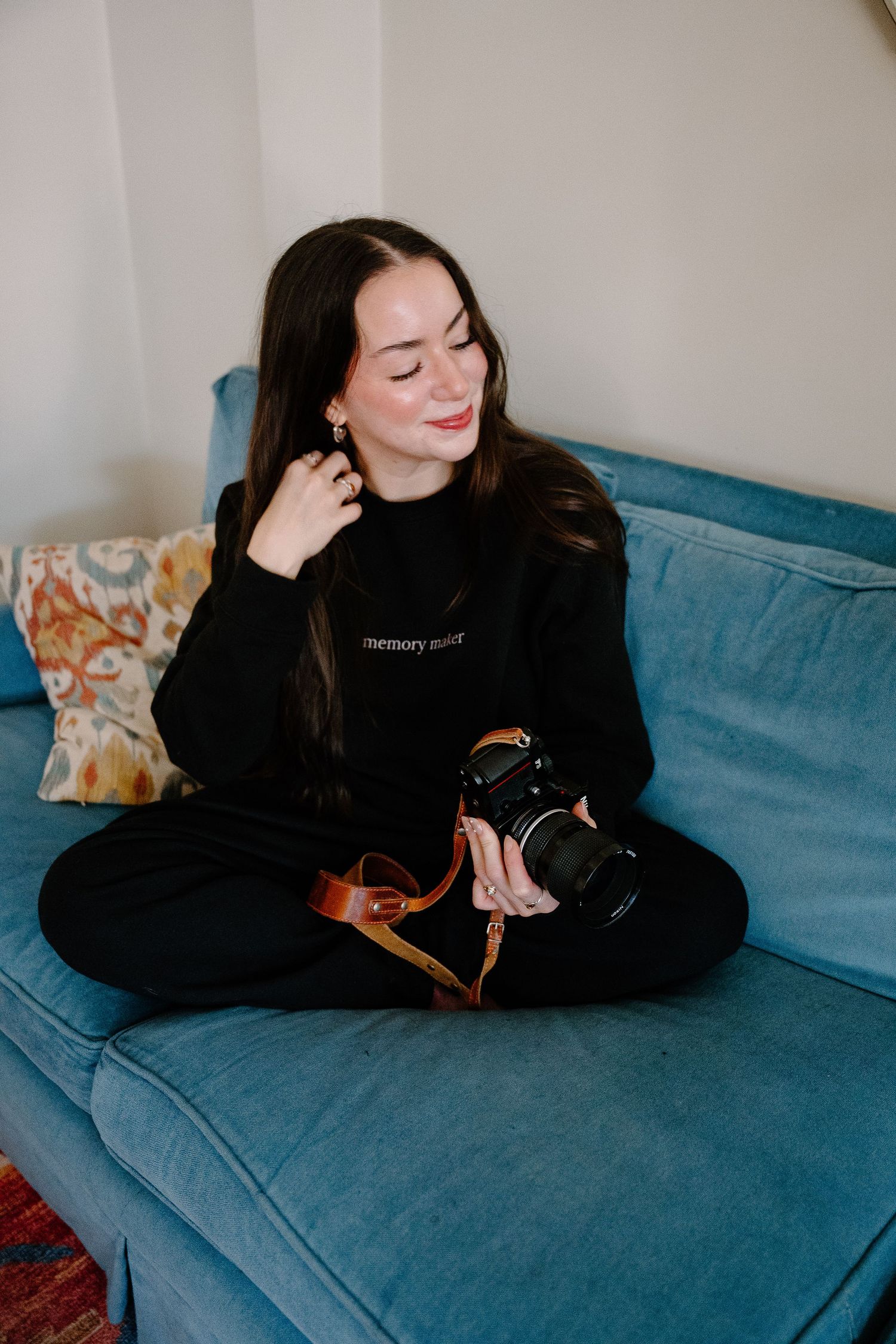 PHOTOGRAPHER in black sweatshirt sitting cross-legged on blue sofa, smiling while holding a DSLR camera with brown strap.