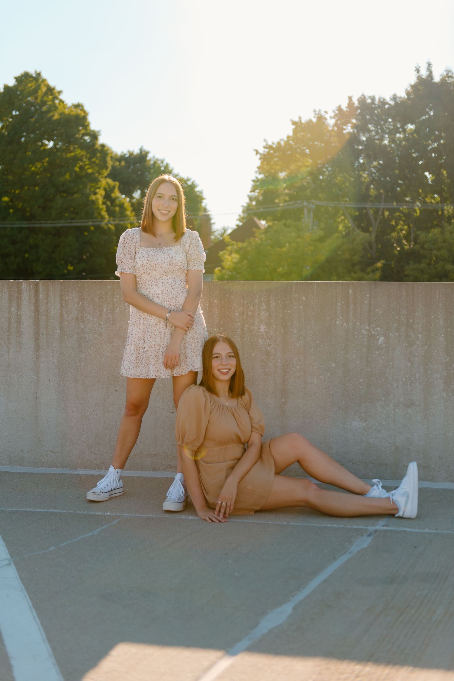 Two friends pose playfully in a parking lot during golden hour sunset, wearing casual summer outfits.