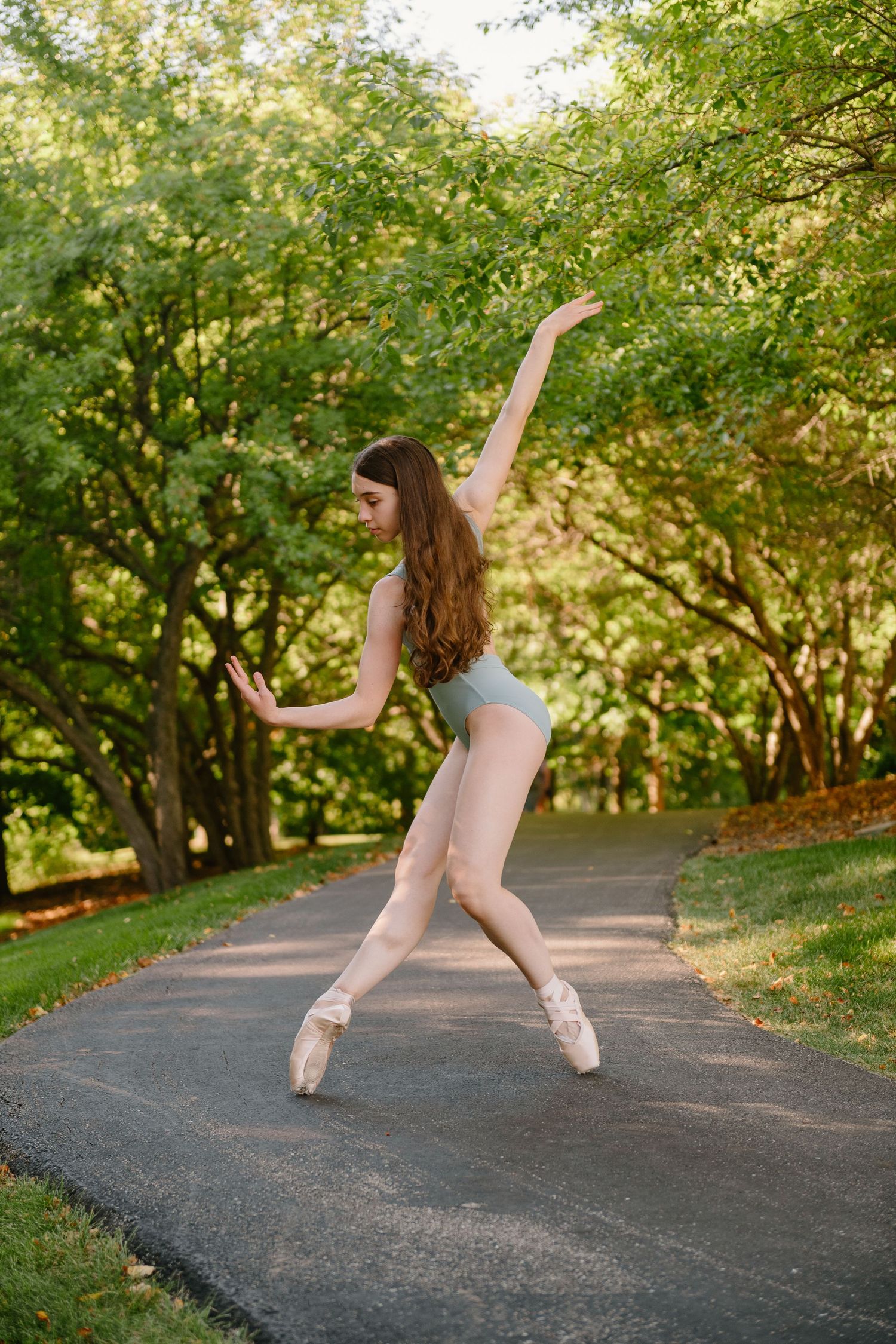 A ballerina in a light blue leotard and pointe shoes performs an elegant pose on an outdoor path surrounded by green trees.