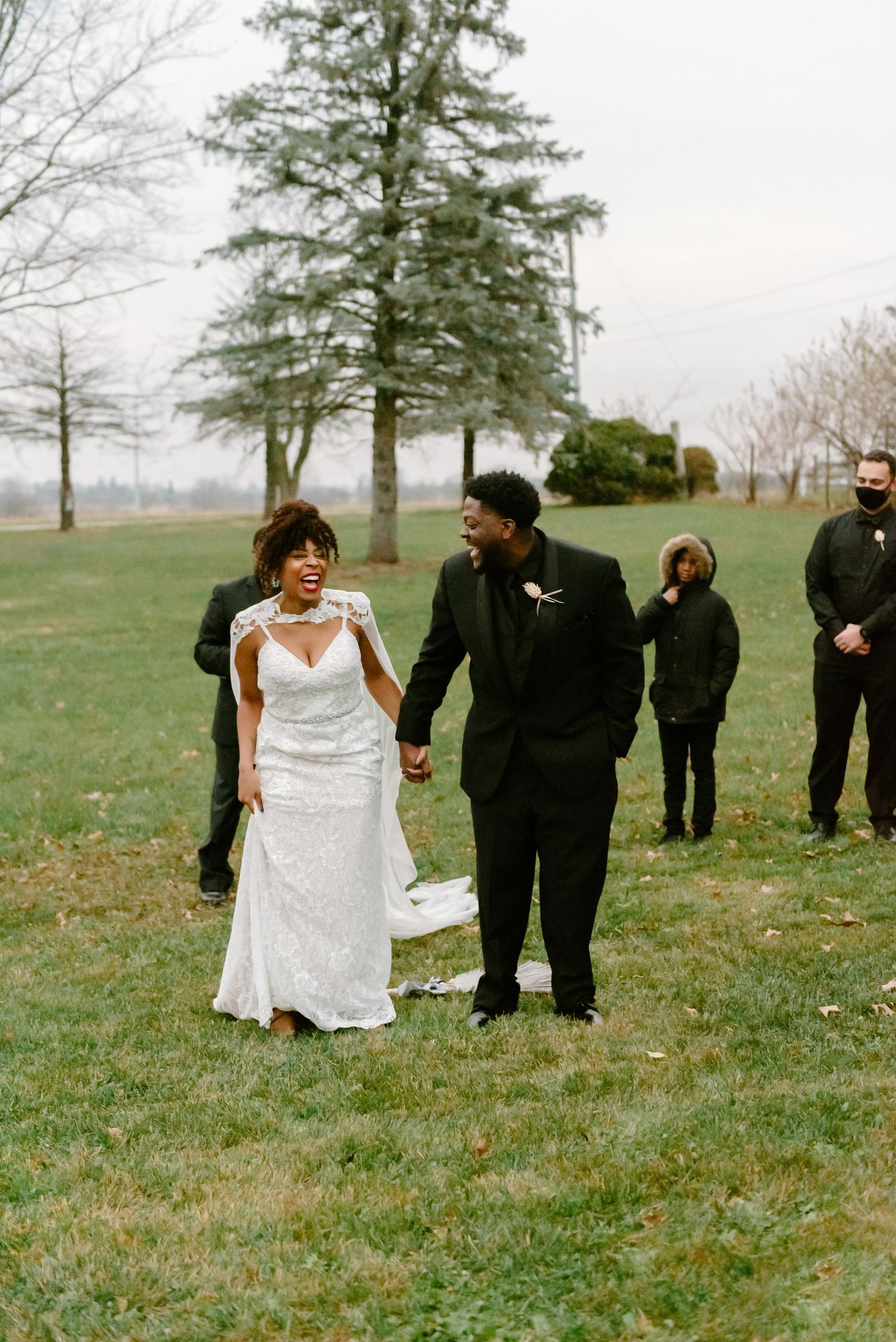 A couple walks hand in hand outdoors at their wedding celebration with guests and tall pine trees in the background.