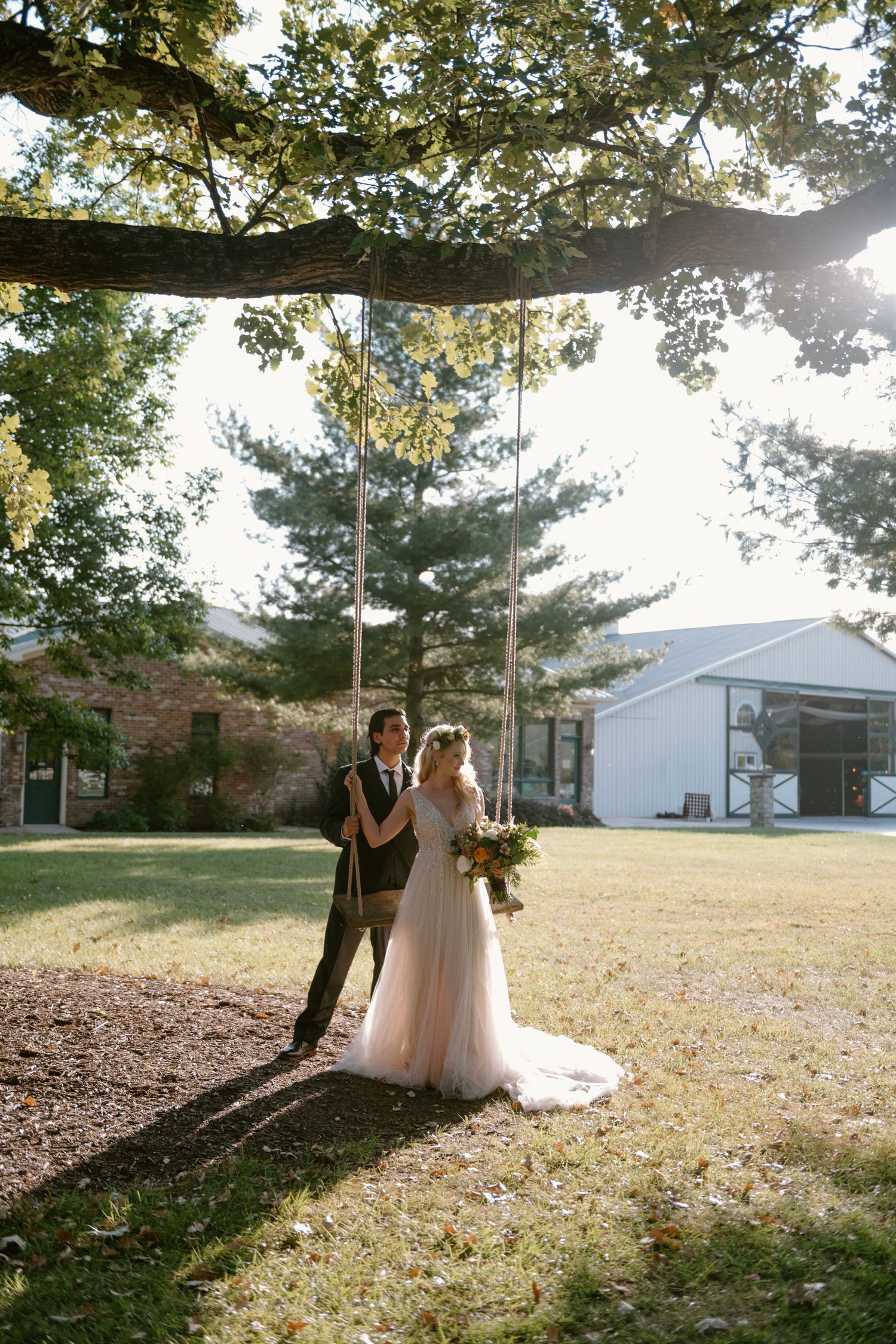 Bride and groom standing by a rope swing under a large oak tree on a sunny day at an outdoor wedding venue.