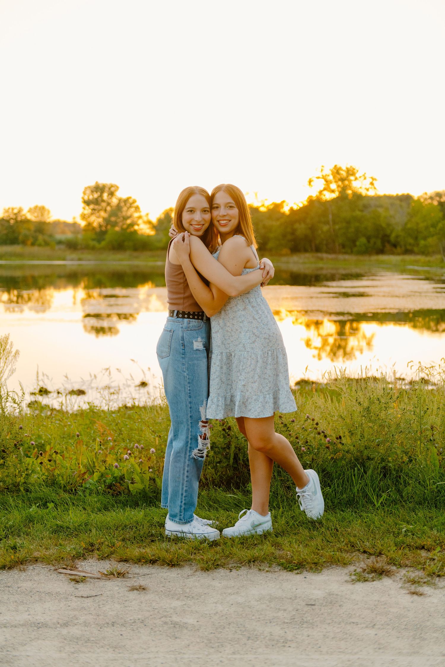 Two friends share a warm embrace by a lake during golden hour sunset with reflections in the water and trees in background.