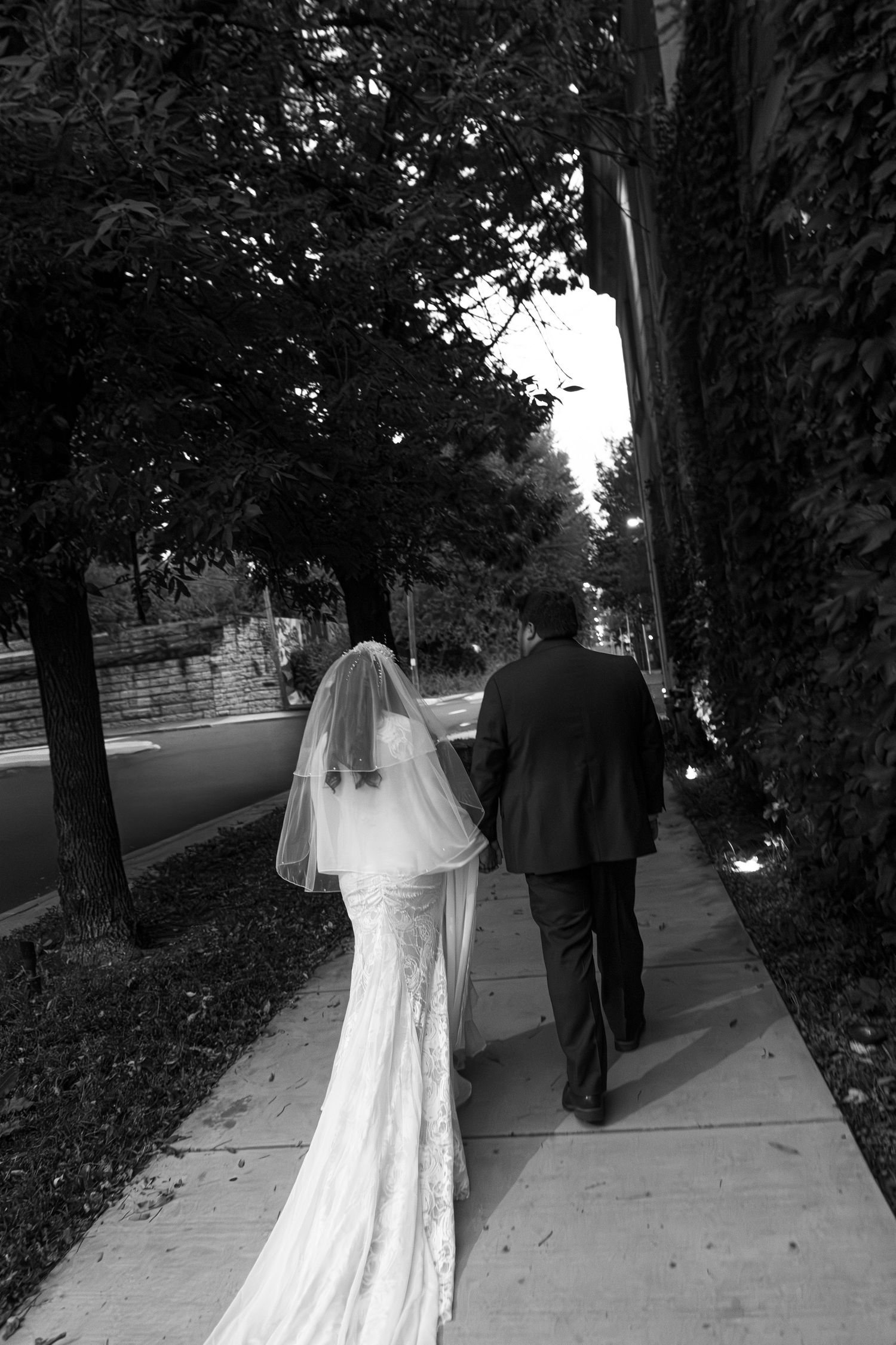 A couple in wedding attire walks down a tree-lined sidewalk in black and white.