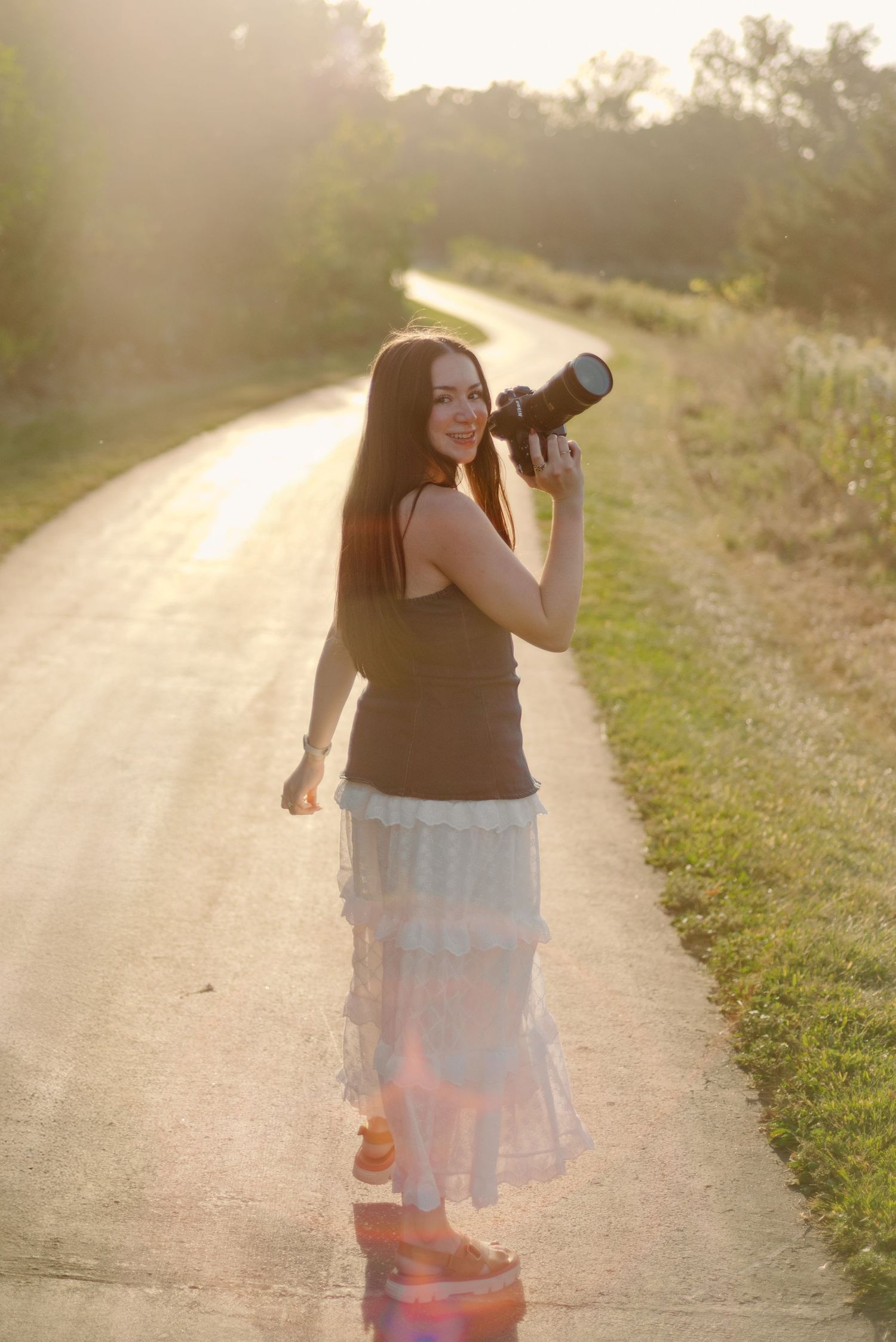 Young PHOTOGRAPHER woman with long dark hair holds a DSLR camera on a sunny rural road, wearing a white lace skirt and olive tank top.